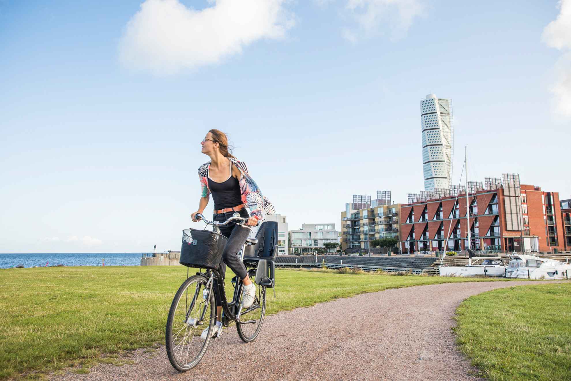 Une femme fait du vélo sur un chemin, derrière on voit un quartier résidentiel au bord de la mer et au milieu un gratte-ciel.
