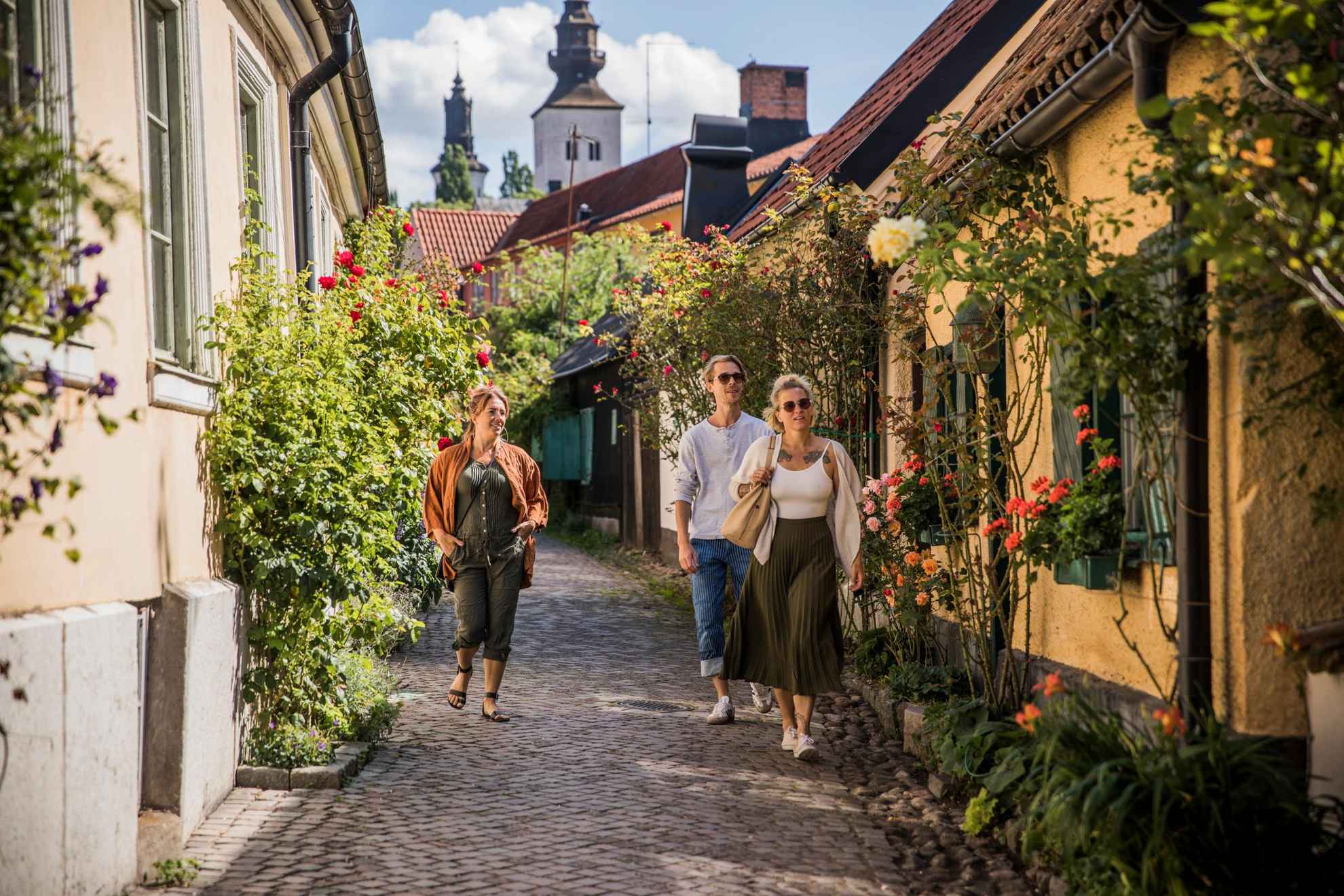 Une homme et deux femmes se promènent dans les rues pavées de Visby. des fleurs fleurissent sur les pans des maisons.