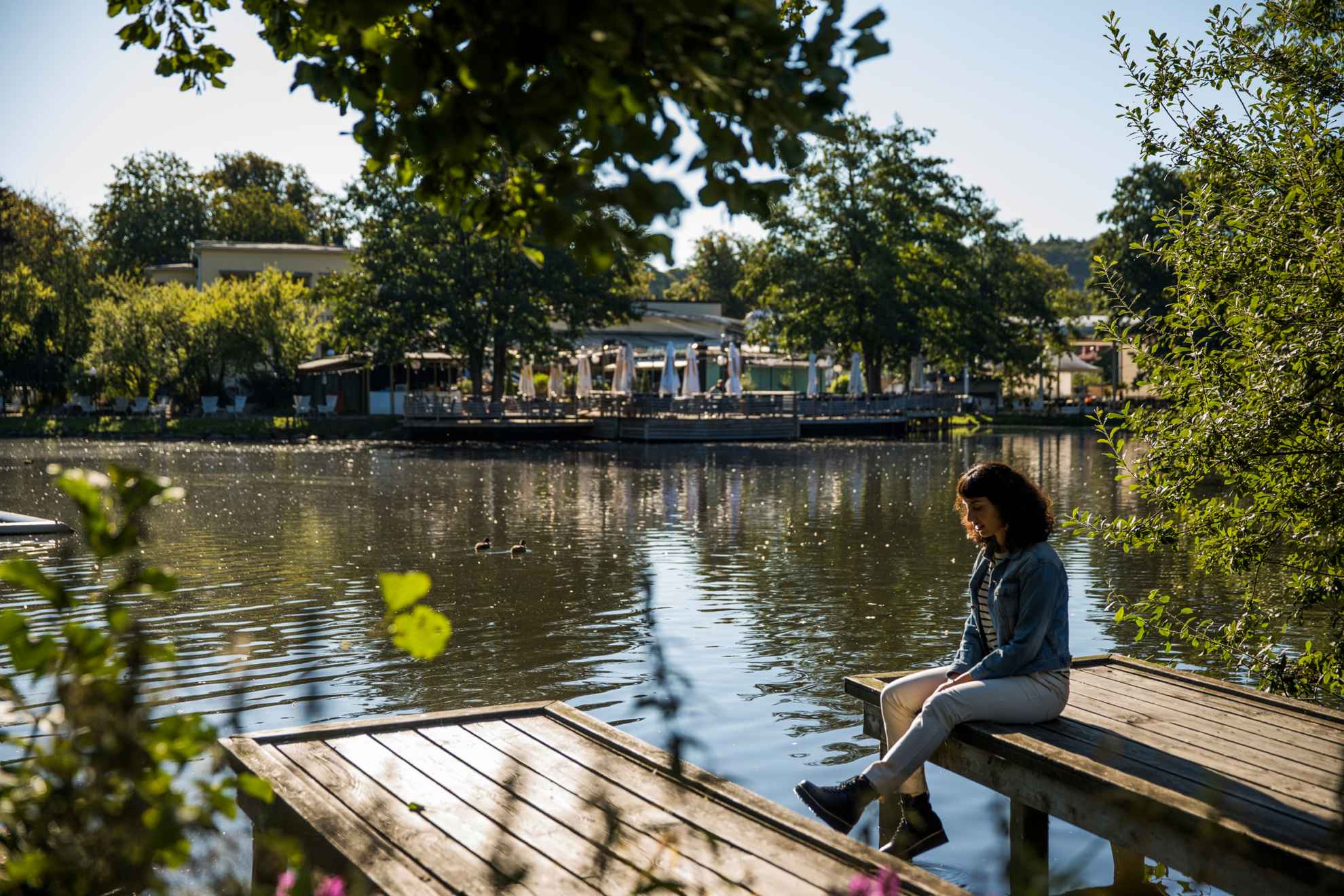Une femme assise sur une jetée près d'un étang entouré de verdure dans un parc. À l'arrière-plan, on aperçoit un restaurant en plein air.