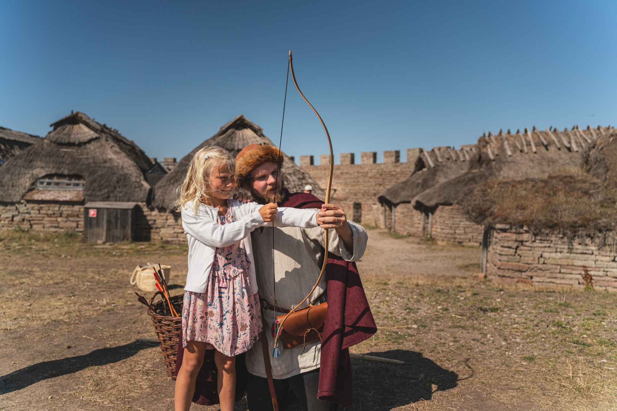 Child tries bow and arrow at Eketorp Fortress with man in period-appropriate clothing.