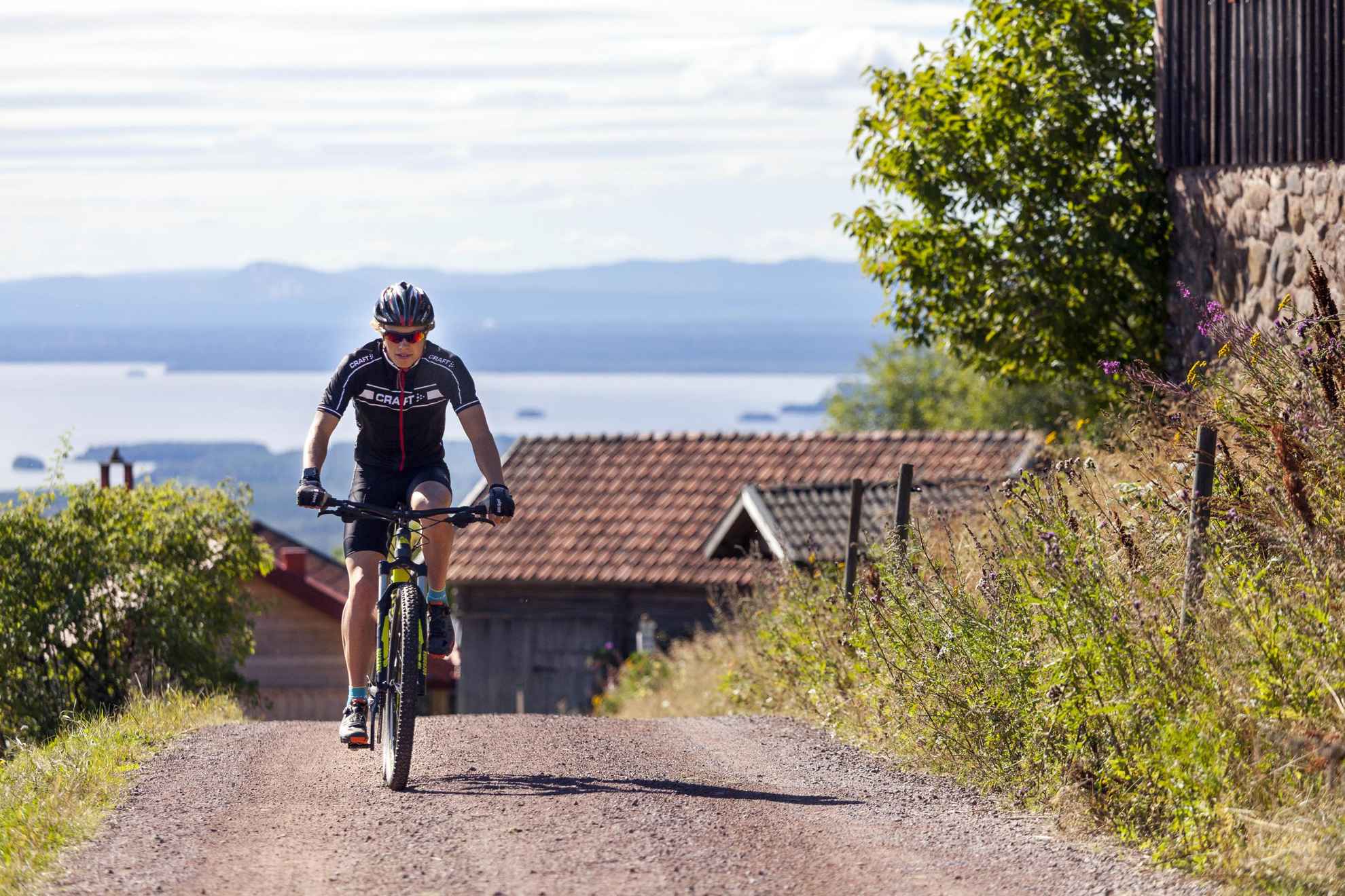 Un cycliste gravissant une colline, avec des maisons en bois, des collines et la mer en arrière-plan.