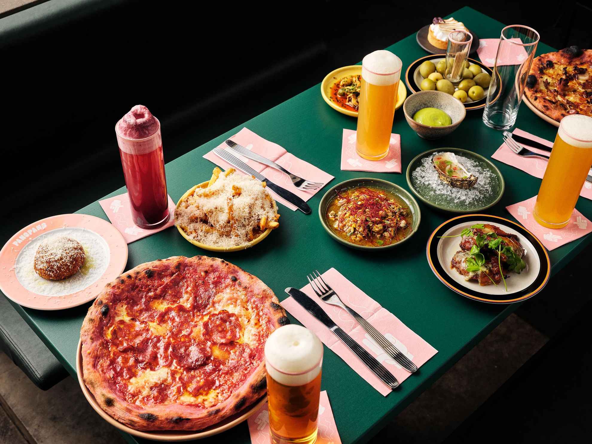 Une table verte sur laquelle sont disposés des couverts, des verres remplis de bière et des assiettes de nourriture avec par exemple des pizzas, des frites, des huitres et des olives.