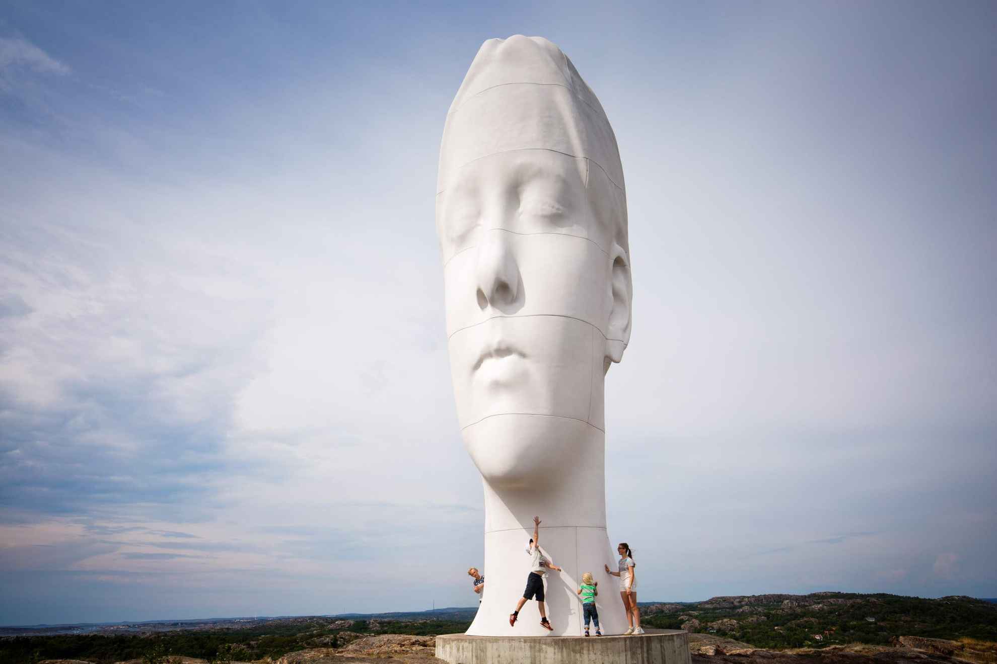 Une grande statue en forme de tête humaine est debout en plein milieu de la nature, avec juste à côté, quatre autres personnes debout.