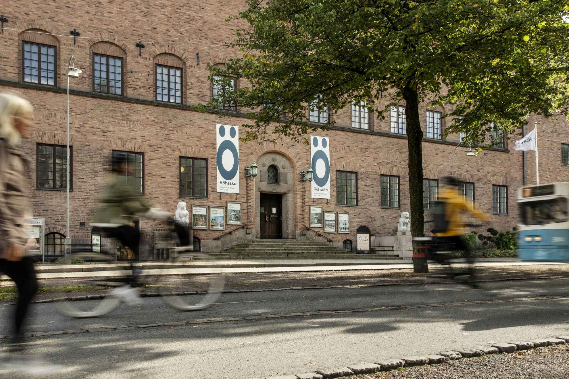Deux personnes à vélo et une personne à pied sont au premier plan en flou devant l'entrée et la façade du musée Röhsska à Göteborg. Le bâtiment est fait de briques et de chaque côté de l'entrée il y a des statues de pierre blanche avec des figures ressemblant un peu à des lions.