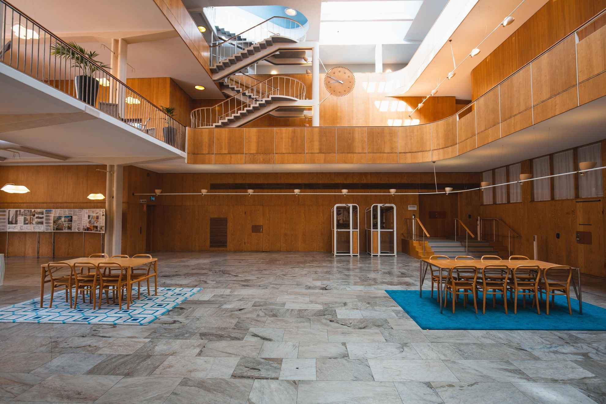 A l'intérieur de l'hôtel de ville on voit une salle avec des murs en bois et un sol en pierres. Deux ensembles de tables et chaises en bois sont situés à gauche et à droite et se trouvent sur un tapis bleu et un tapis à motifs. Au niveau des étages supérieurs, on aperçoit des balcons et des escaliers.