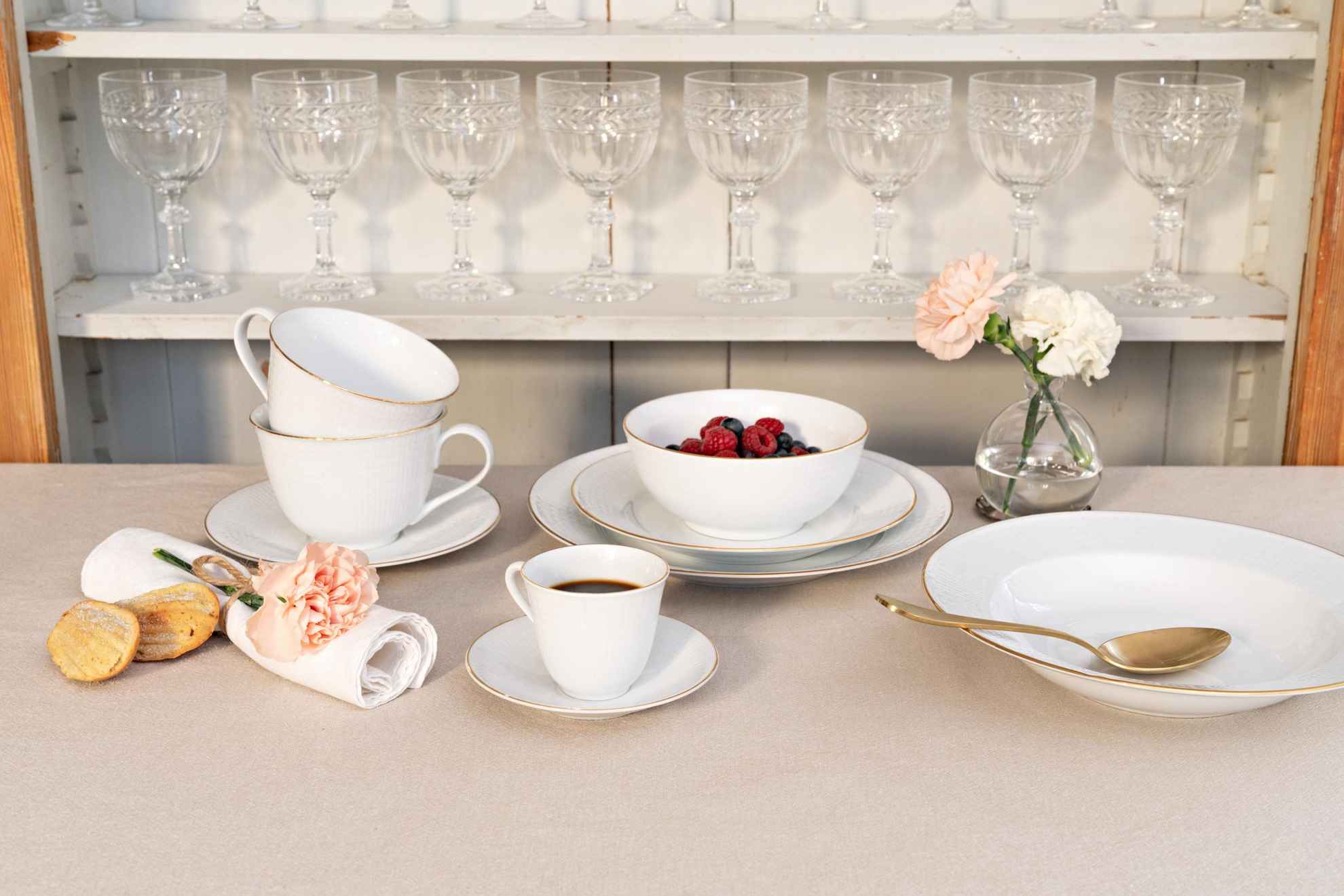 Un service de table appelé "Swedish Grace" est posé sur une table. Il y a des assiettes de différentes tailles, un bol rempli des baies, deux tasses à thé et une tasse de café. Derrière il y a une étagère rempli avec plusieurs verres à vin.