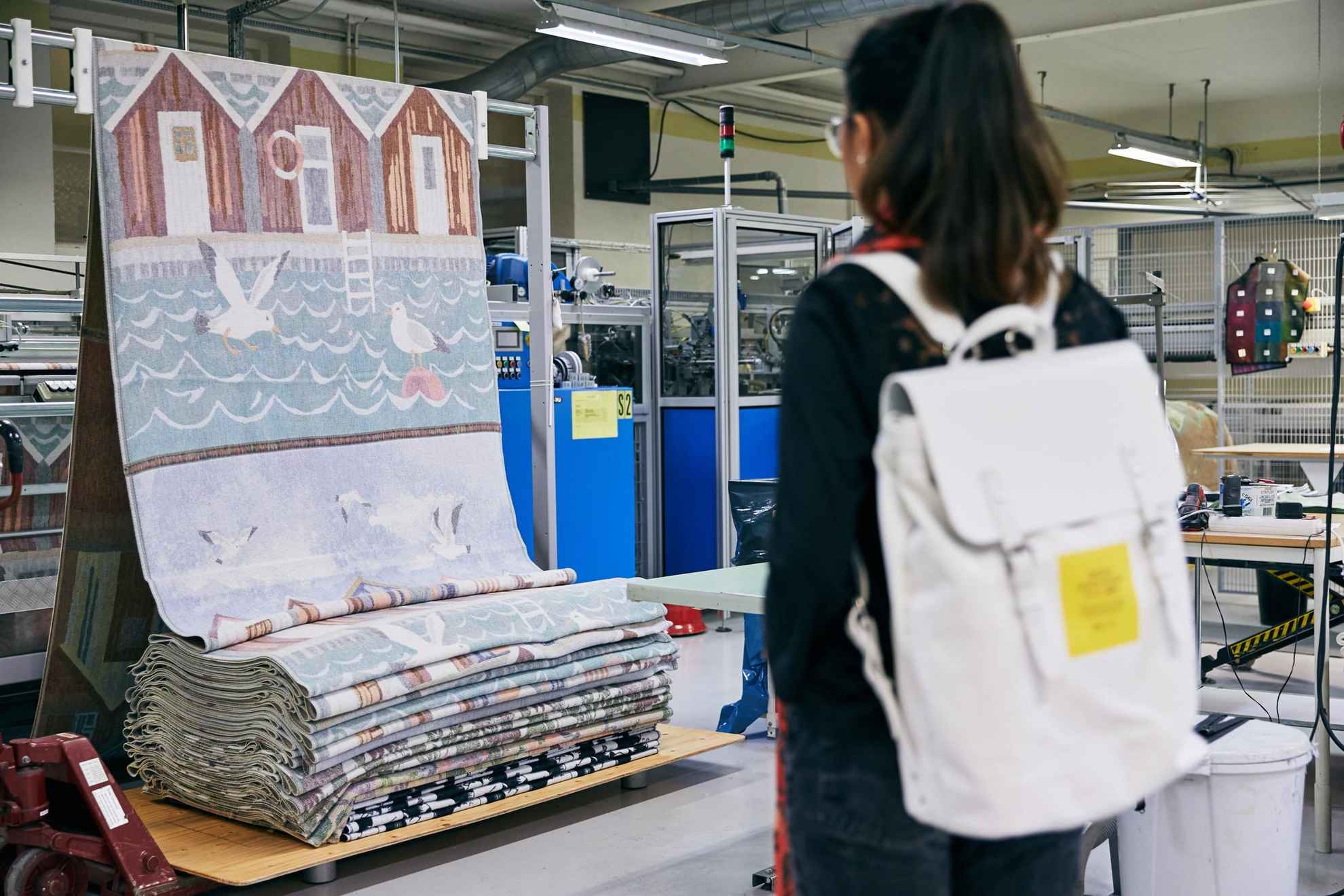 Une femme avec un sac à dos visite une usine de tissage et regarde un tissu avec des cabanes rouges et des oiseaux. Le tissu est suspendu sur un support.