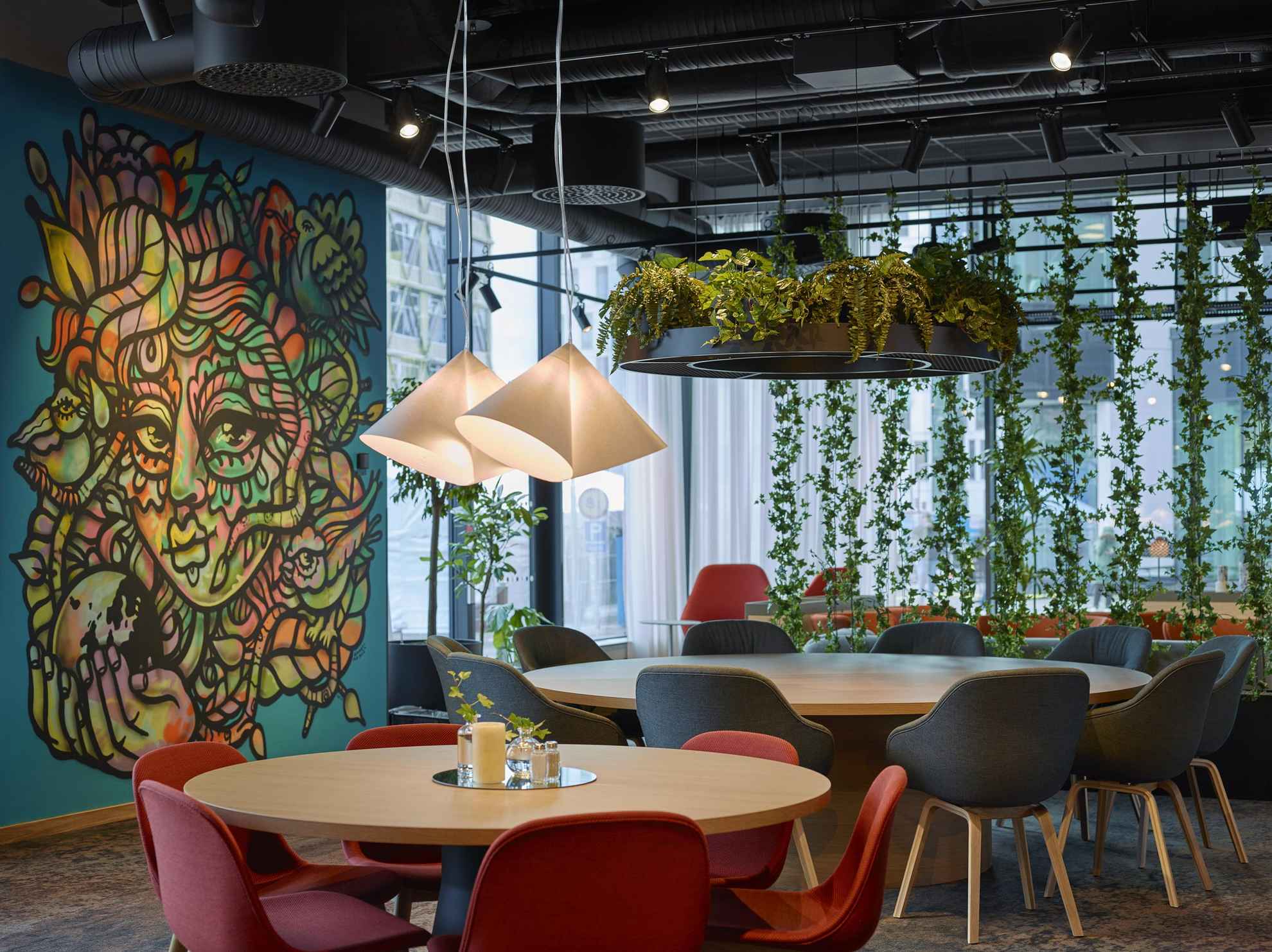 Une pièce avec des tables rondes en bois et des chaises noires et rouges. Des plantes sont suspendues au plafond et sur la gauche de la photo une oeuvre a été peinte sur un mur bleu.