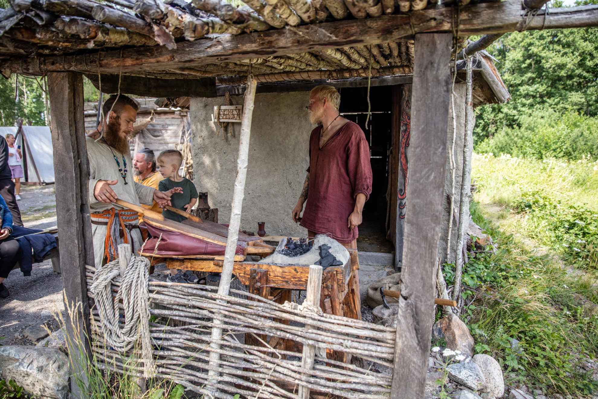 Deux enfants regardent deux hommes, vêtus de vêtements typiquement vikings, travailler autour de l'âtre.