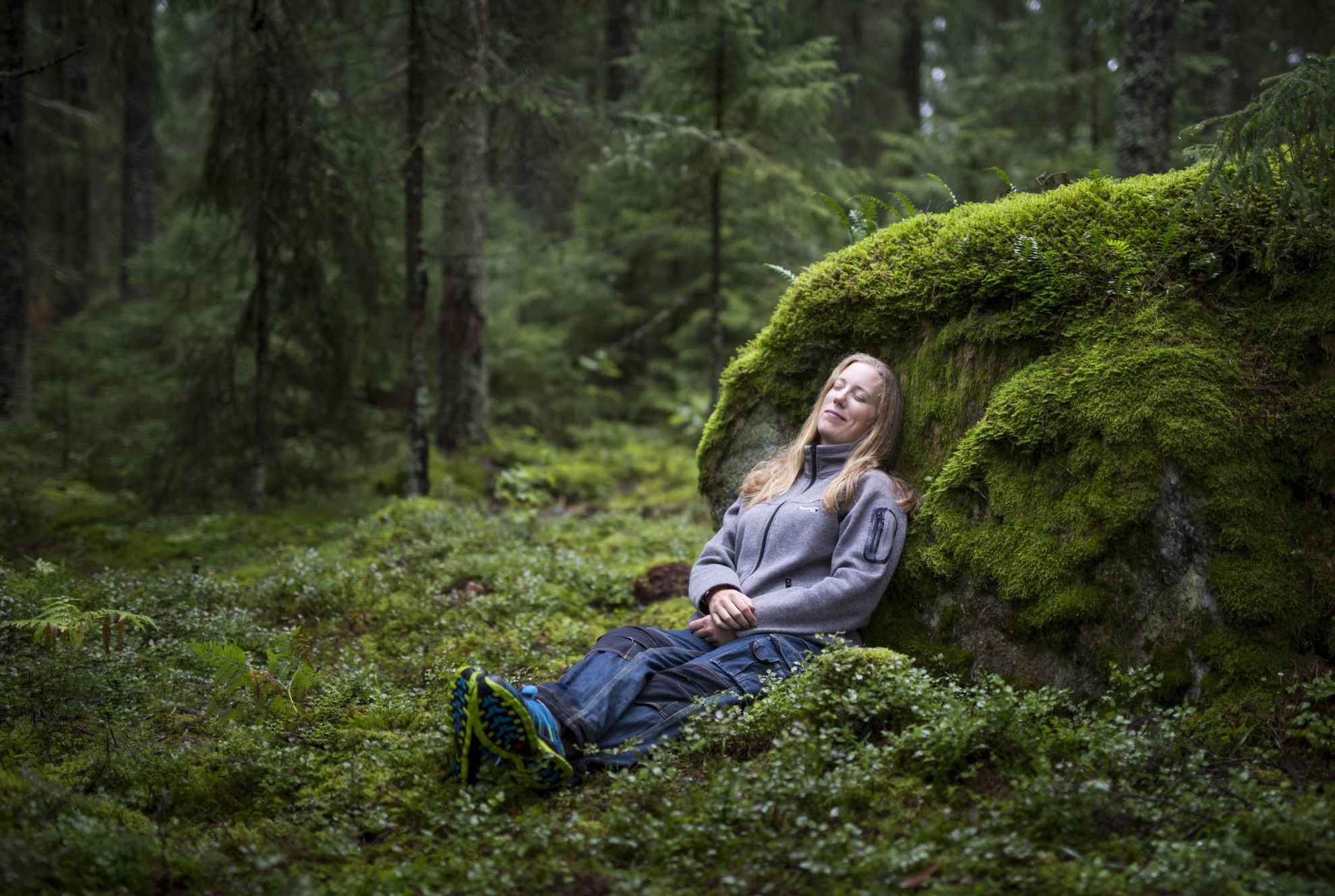 Une femme est assise par terre dans une forêt et s'appuie contre un rocher recouvert de mousse.
