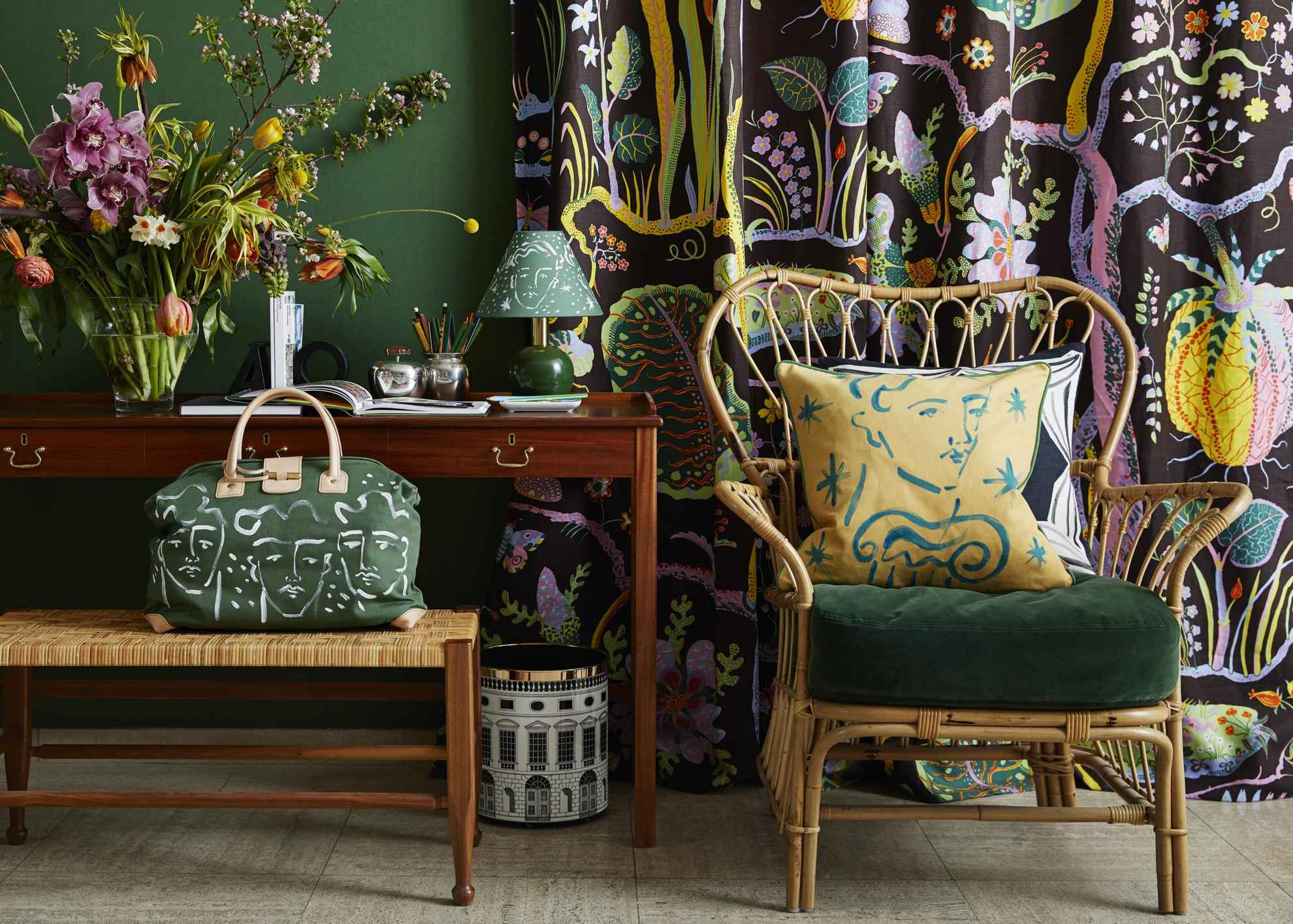 Des fleurs sont disposées dans un vase avec une lampe sur une table à côté d'une chaise avec un coussin. Devant la table se trouve un sac posé sur un tabouret  Derrière la chaise se trouve un rideau à motifs colorés.