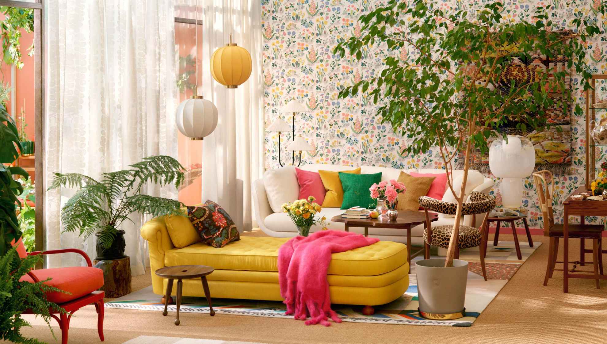 Un salon avec un canapé beige avec des coussins colorés à l'arrière et une chaise longue jaune au centre de la pièce. À droite de la chaise longue se trouve une grande plante verte au sol. Les rideaux blancs transparents laissent entrer la lumière naturelle.