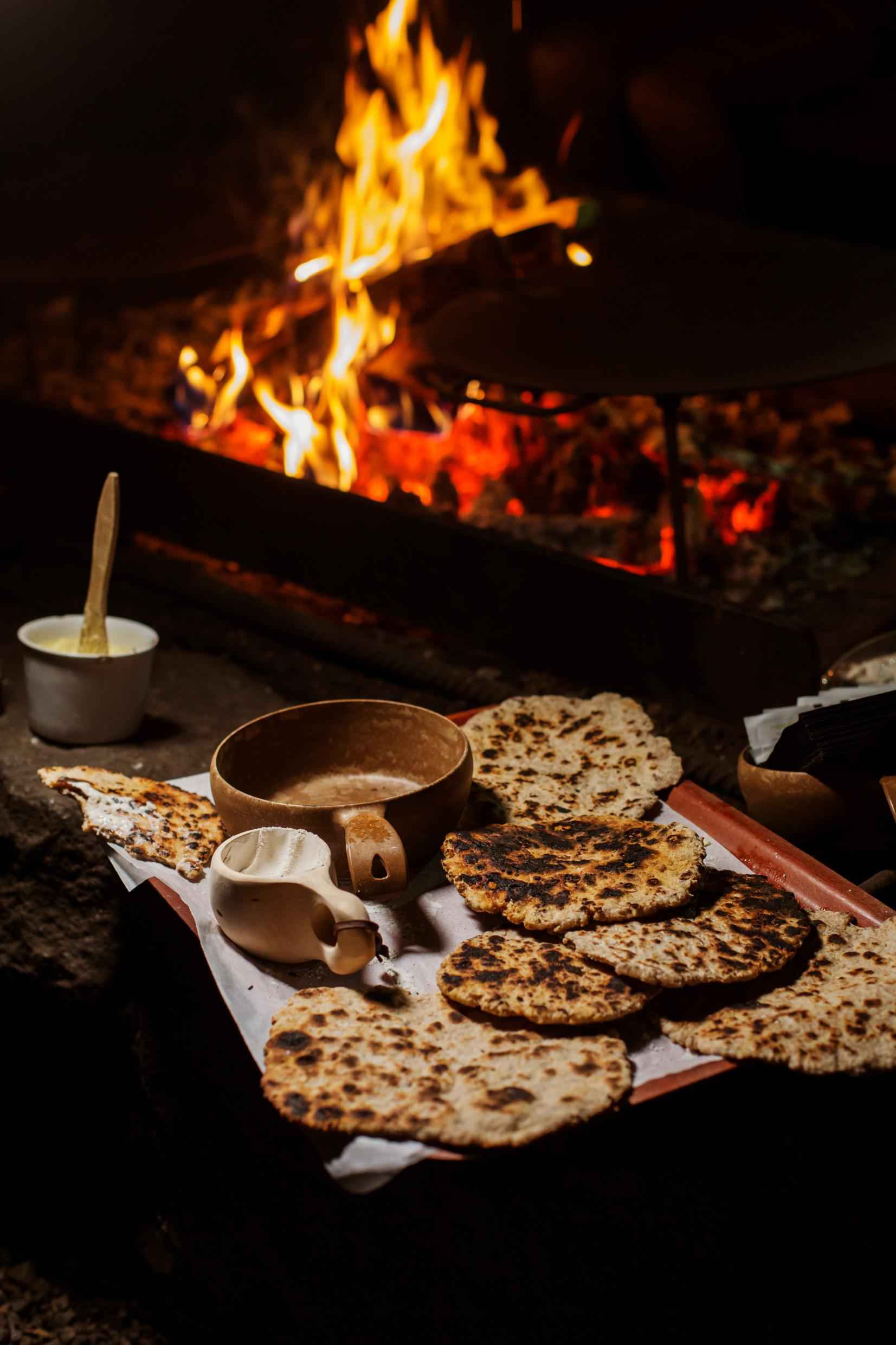 Un plateau avec du pain plat fraîchement cuit et deux tasses en bois. Un pot de beurre est situé à côté du plateau avec un feu de camp en arrière-plan.