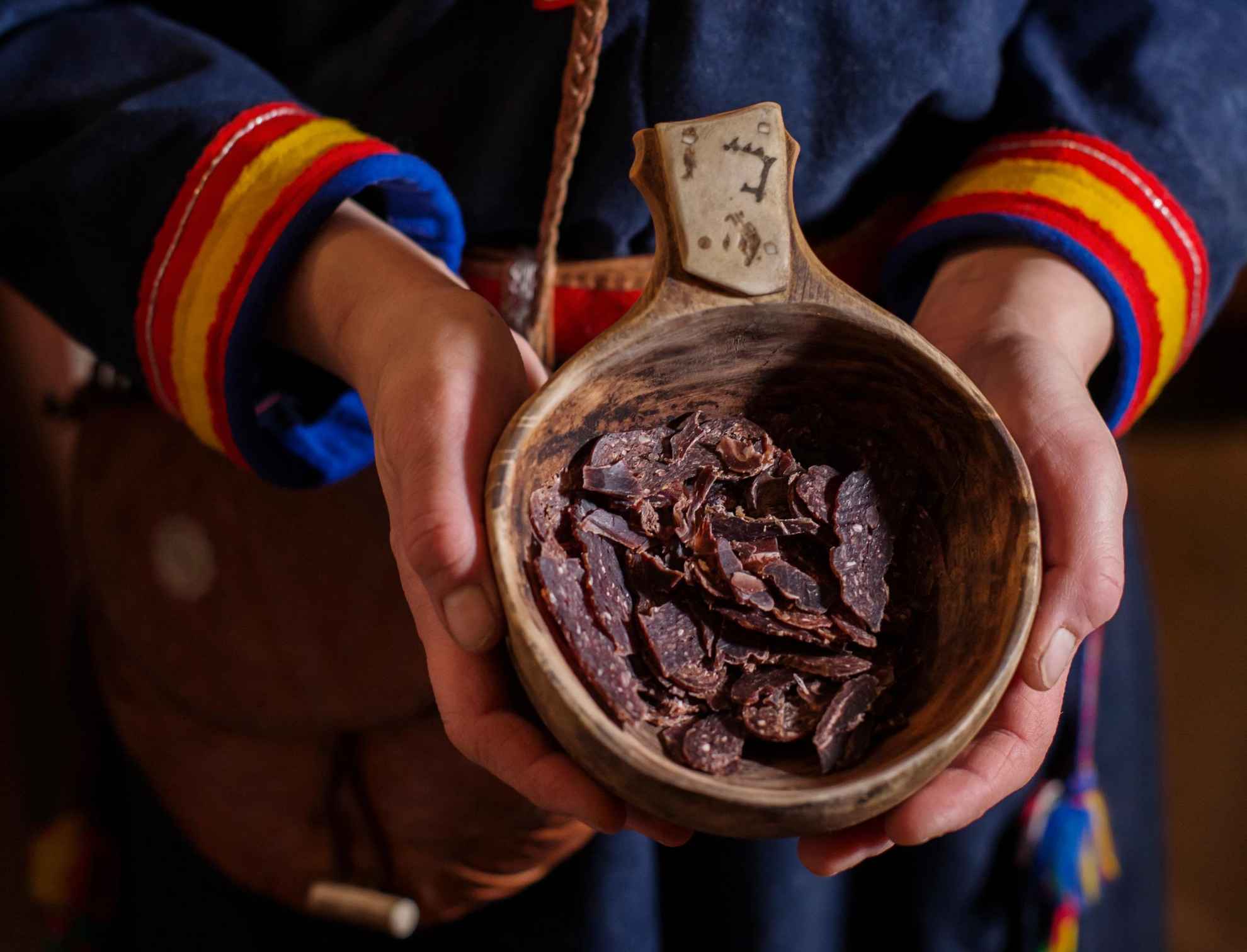 Une personne vêtue de vêtements samis tenant une tasse en bois remplie de morceaux de viande de renne séchée.