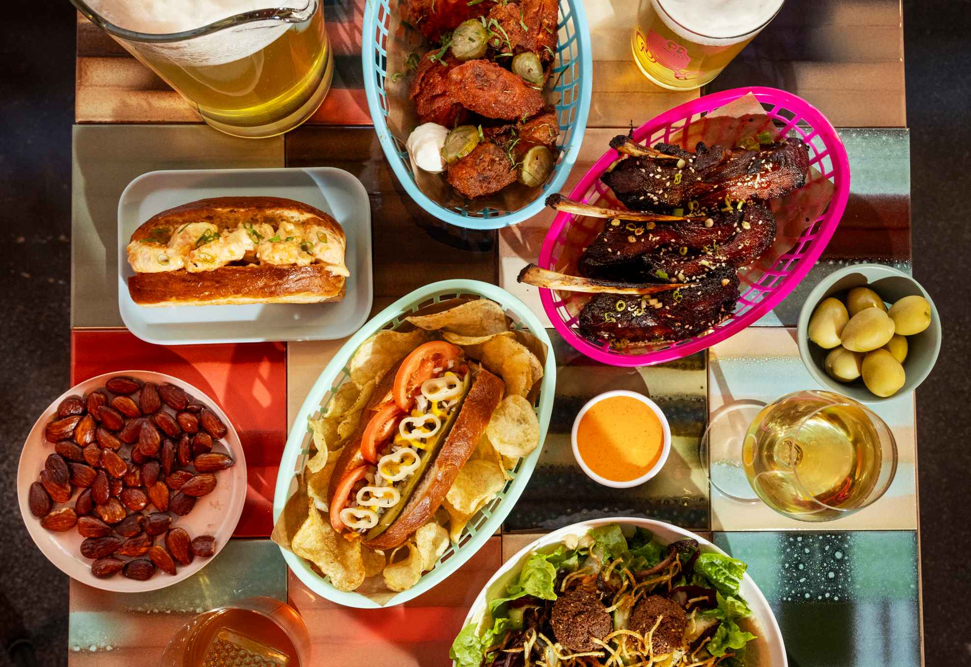 Une table carrée remplie de streetfood colorés dans différents paniers et sur des assiettes, ainsi que des verres de bière et de vin.