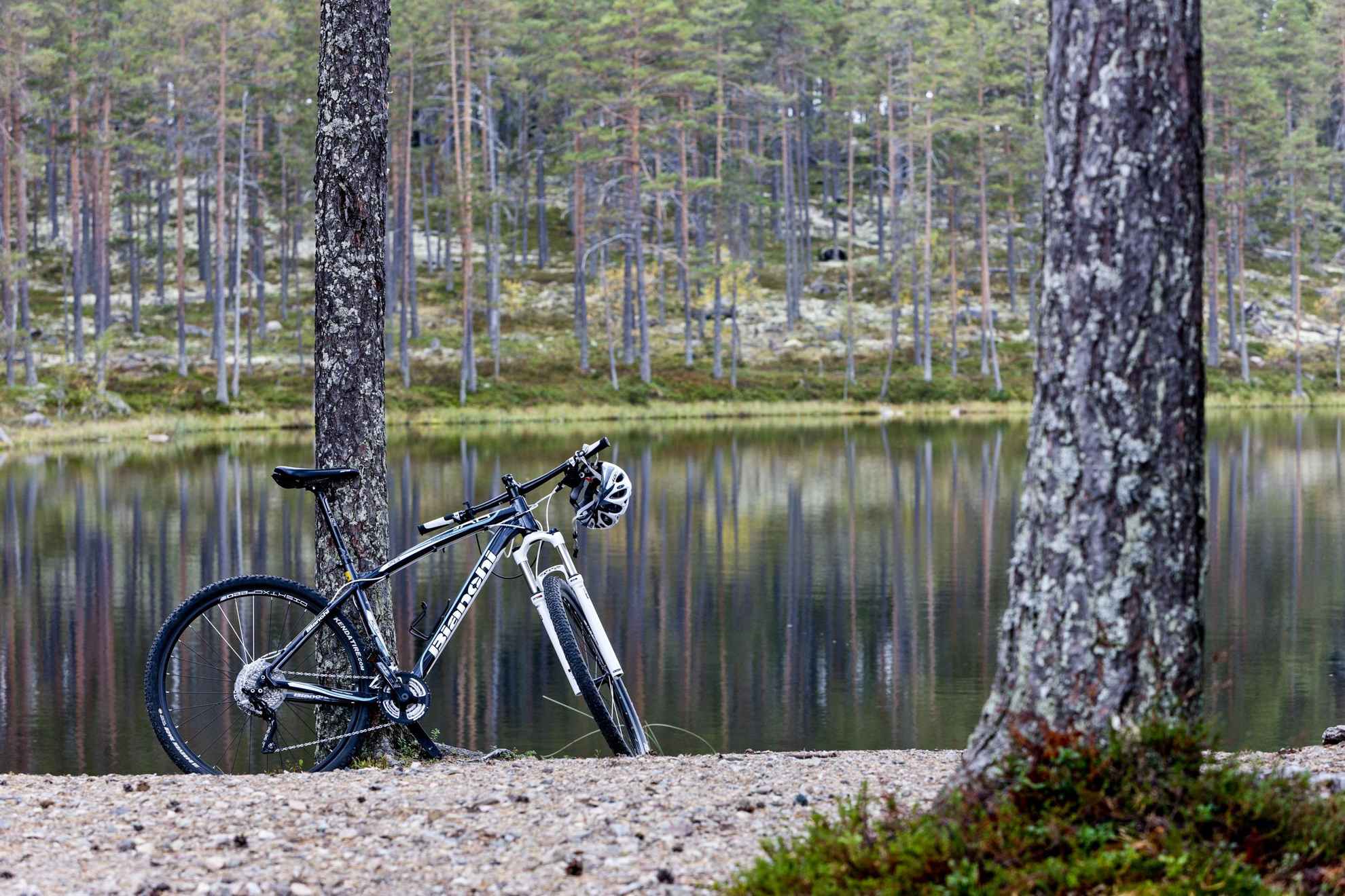 Un vélo est debout avec sa béquille devant un lac dans la forêt et on voit le reflet des arbres dans l'eau.