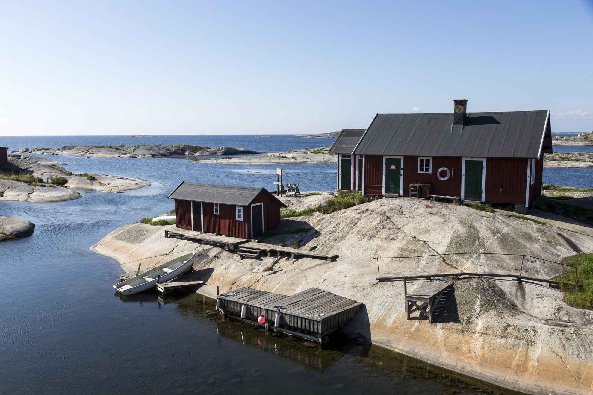 Une maison et un hangar à bateaux avec un ponton en bois se trouvent sur les rochers de l'archipel de Stockholm. Une barque vide est posée sur les rochers et flotte en partie dans l'eau.