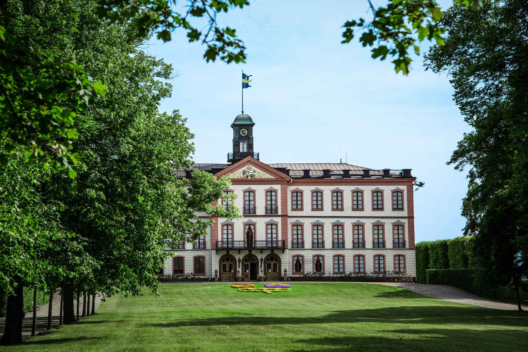 L'extérieur du château de Tullgarn, avec des murs blancs et de grandes fenêtres entourées de détails rouges. Des arbres et une pelouse entourent le bâtiment.