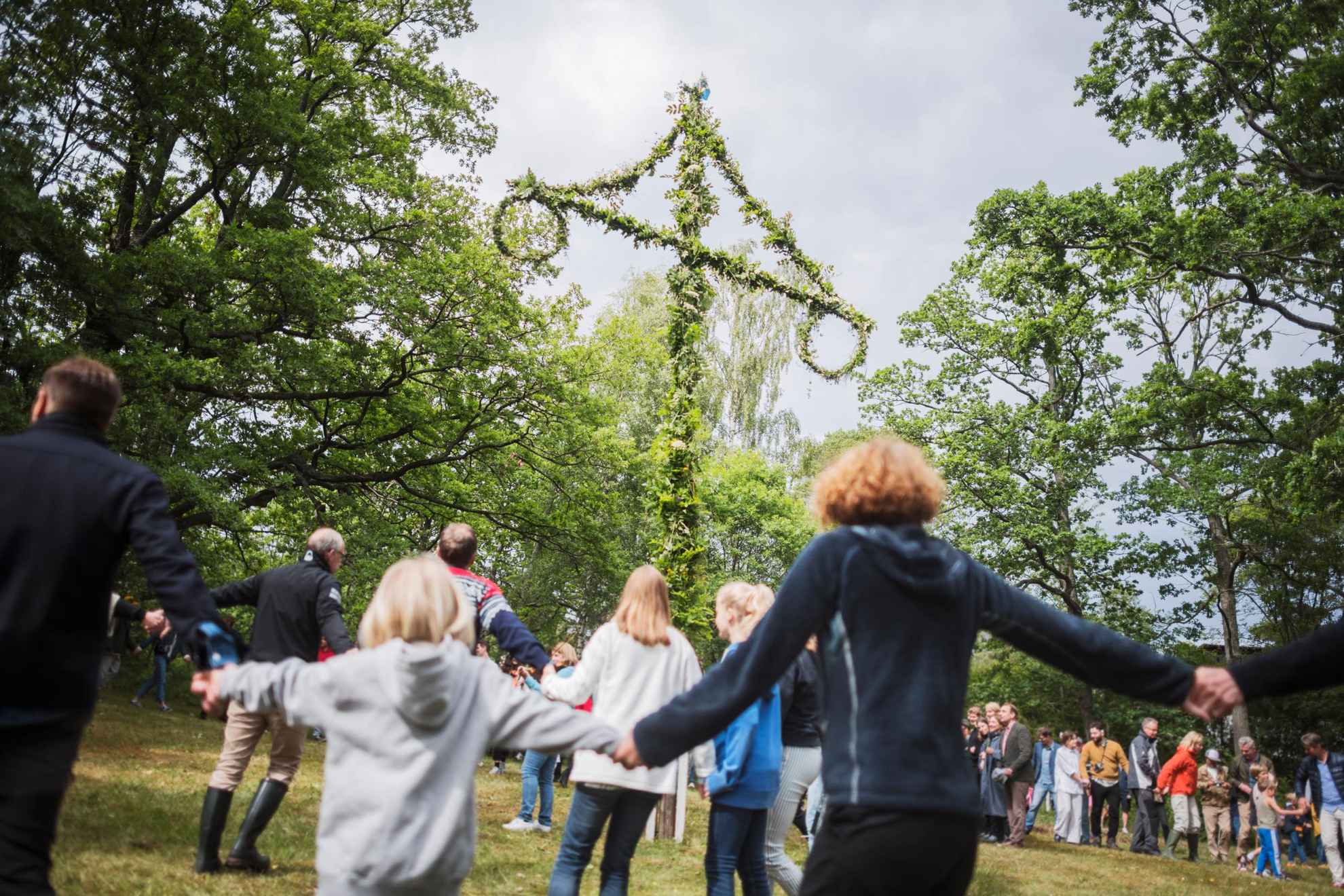 Kids and adults are holding hands and dancing around a midsummer pole. A midsummer pole is a large pole stuck into the ground with a shorter cross bar from which hangs two wreaths.