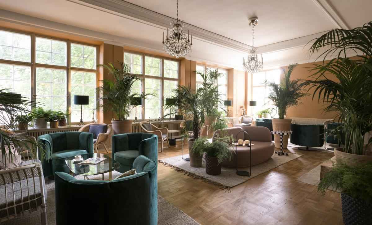 Un grand lounge avec des canapés et des fauteuils. La pièce est décorée avec des plantes, et deux chandeliers au plafond.