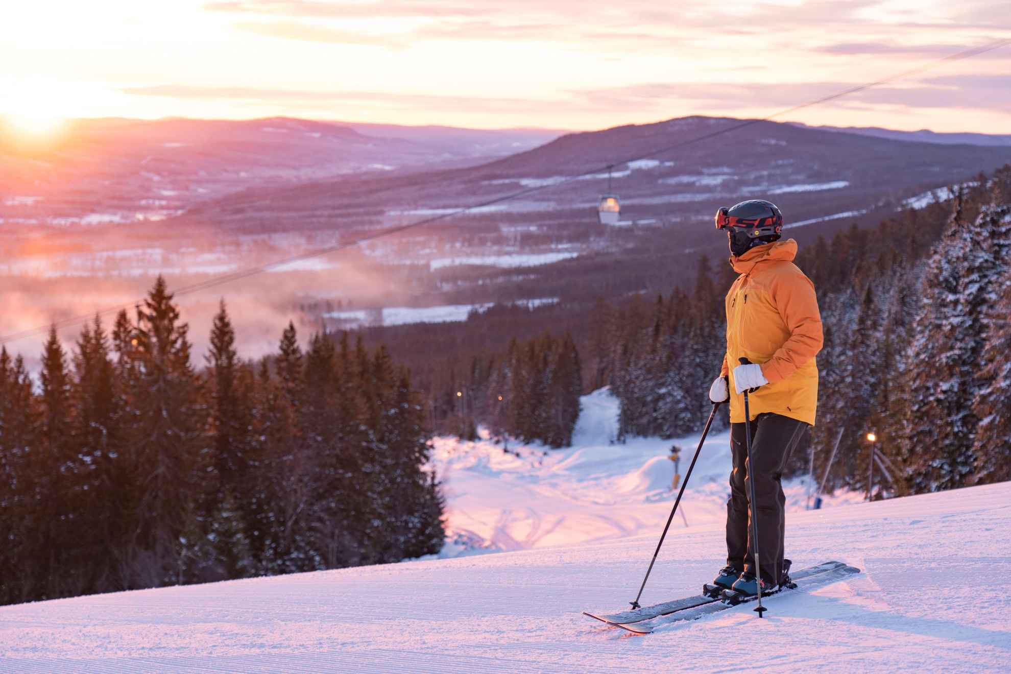 Une personne avec du matériel de ski se tient sur une piste de ski en regardant le coucher du soleil.