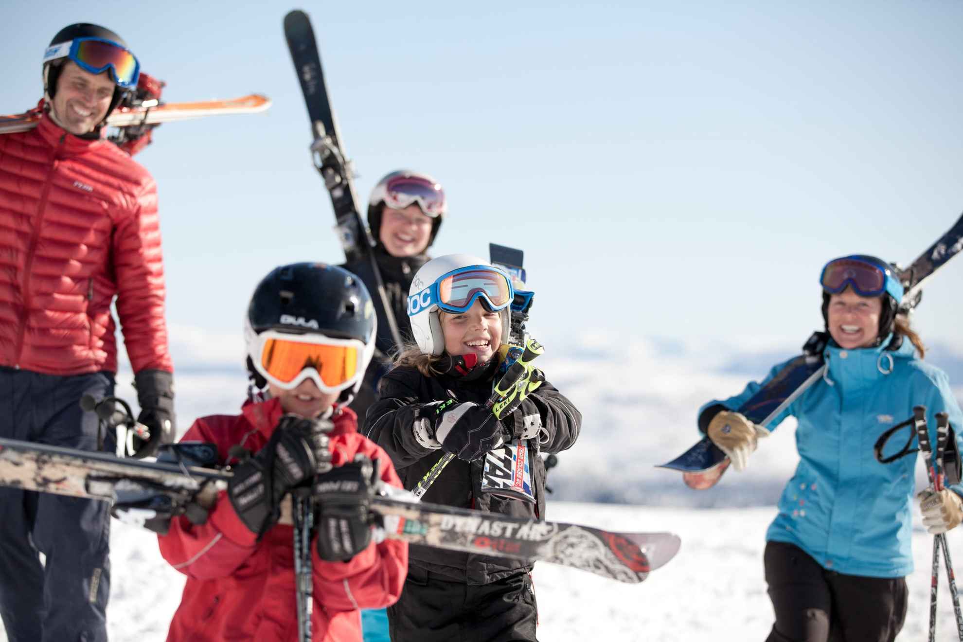 Des enfants et leurs parents portent leur ski dans les montagnes tout en souriant.