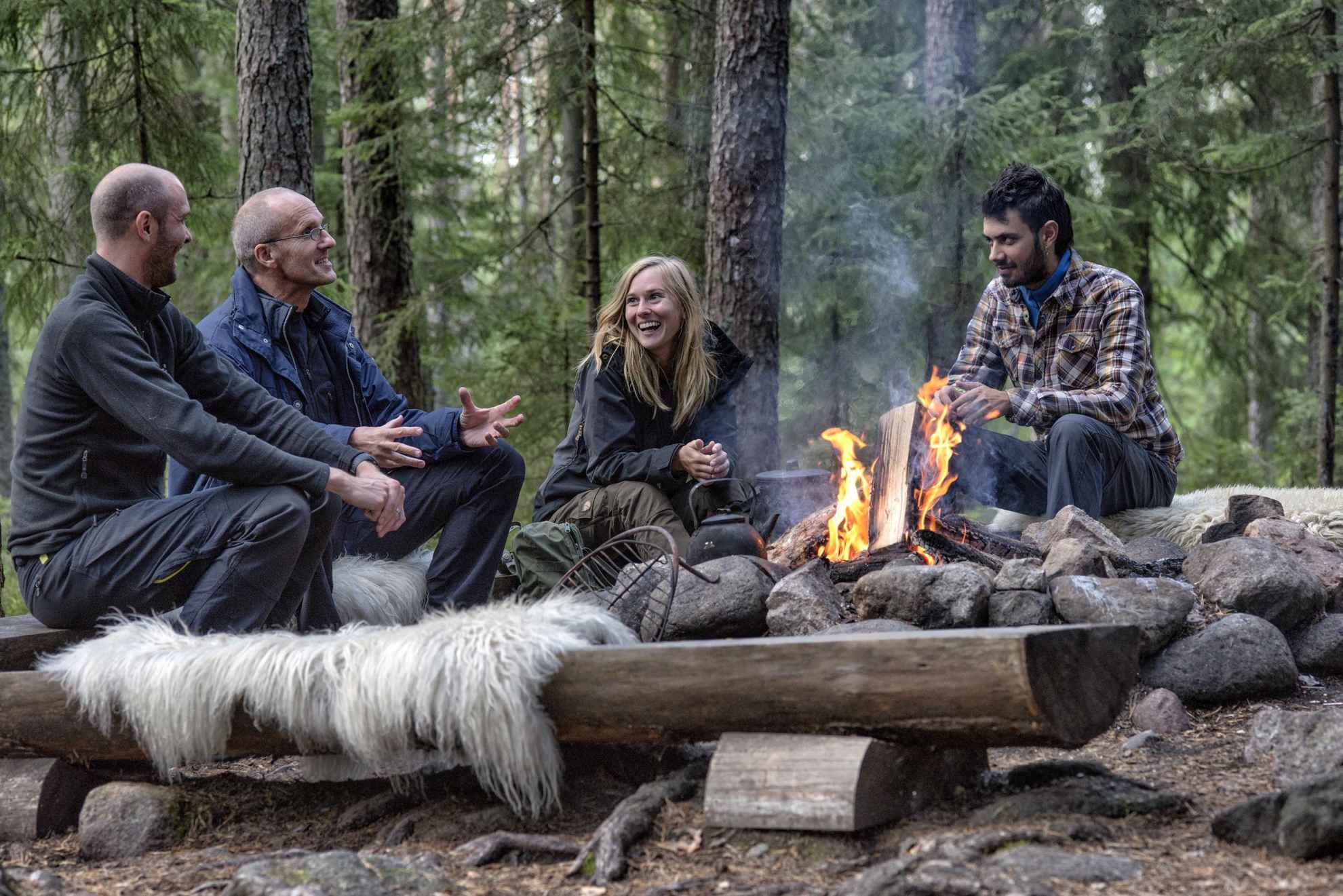 Quatre personnes sont en train de rire, assises autour d'un feu de camp, dans une forêt.