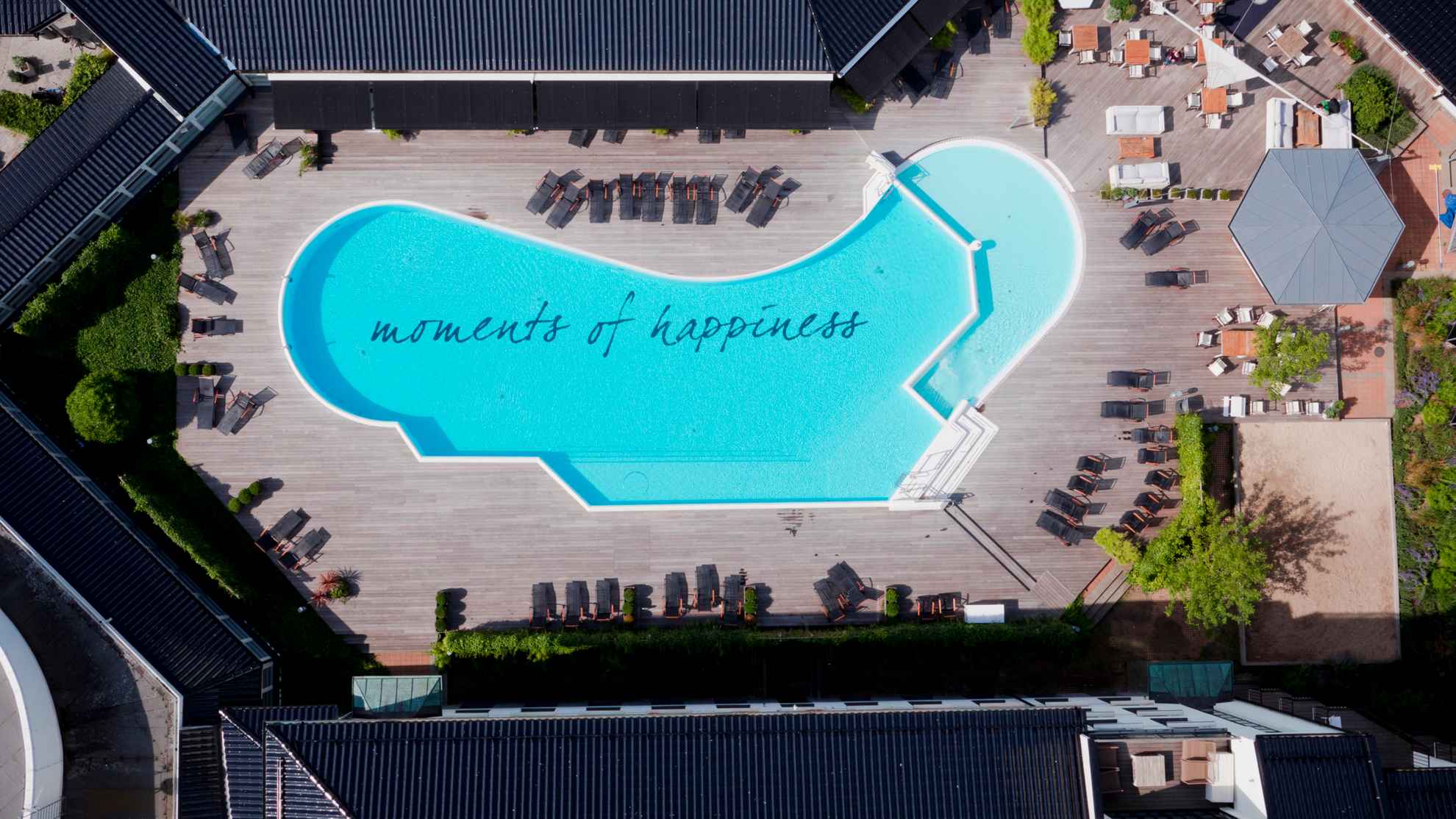 Une piscine extérieure avec l'inscription "moments de bonheur" au fond, entourée d'une terrasse en bois et de chaises longues.