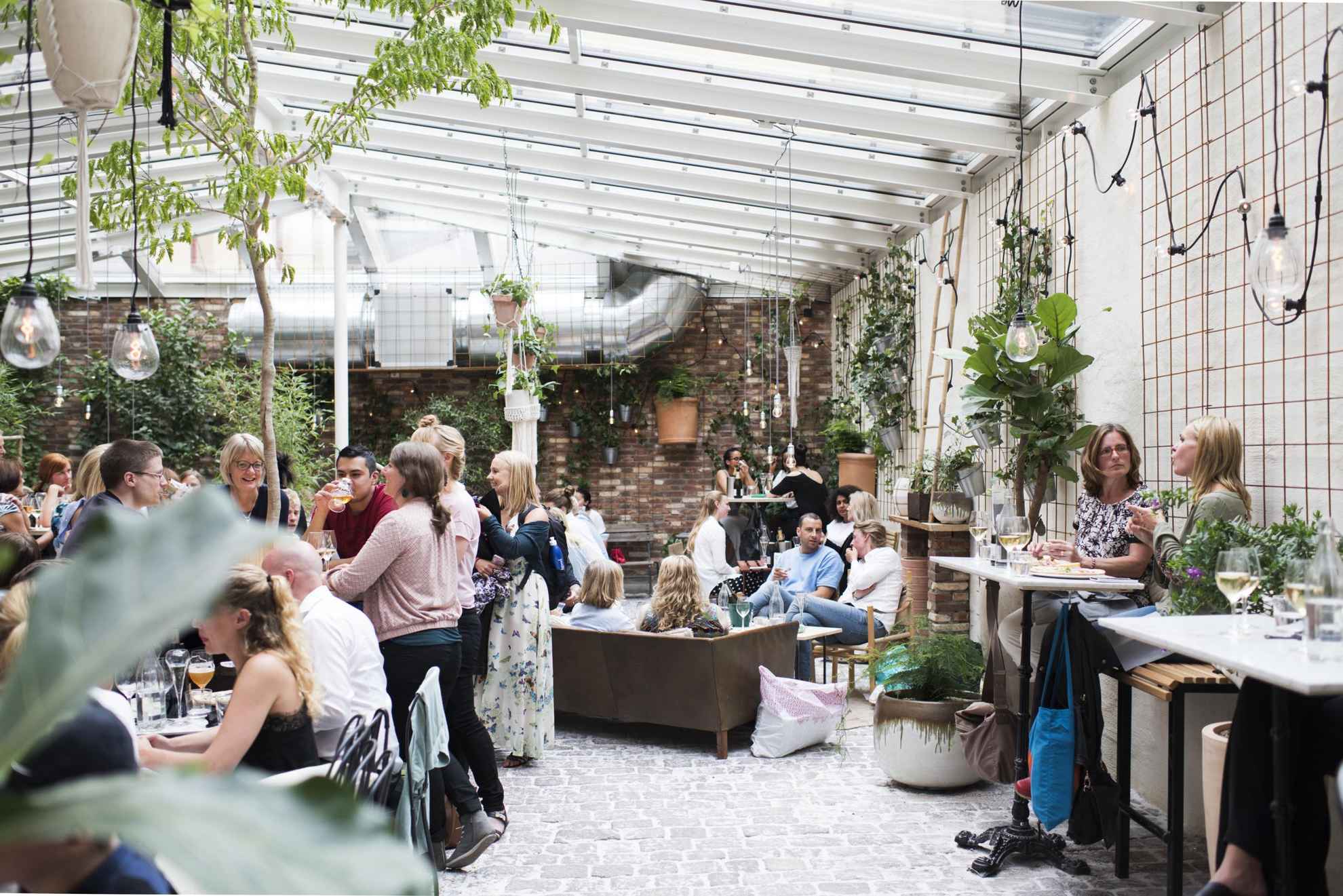Des gens apprécient les boissons et la nourriture dans le café Magasinet de Göteborg. Le sol du café est en pierre, les murs en briques, le plafond de verre et l’intérieur est rempli de plantes vertes.