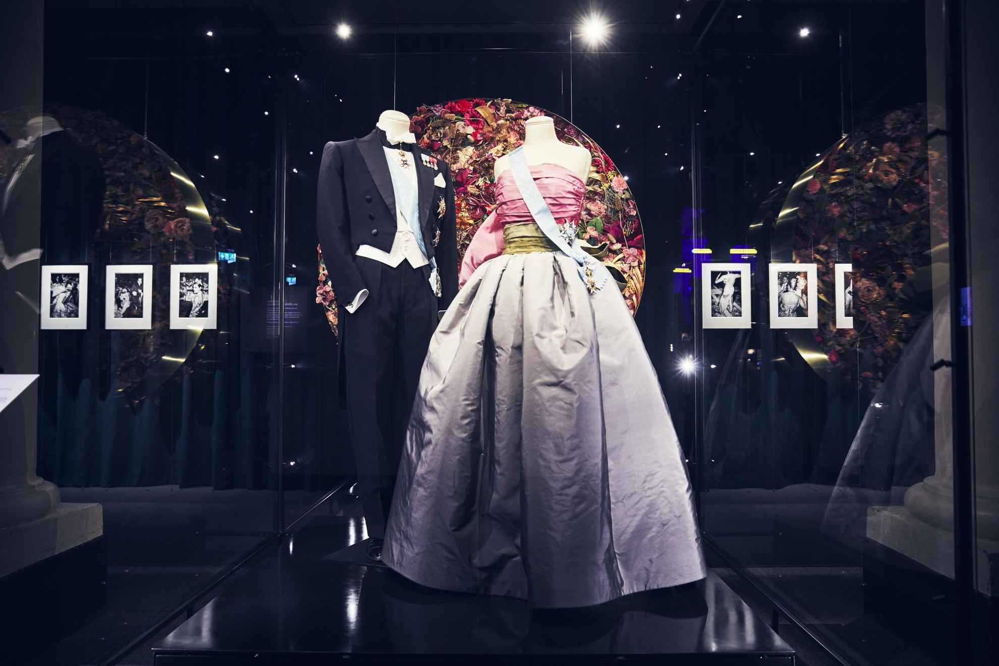 Deux mannequins dans une exposition, l'un vêtu d'un frac et l'autre d'une robe violette et rose. Tous deux portent un ruban bleu.