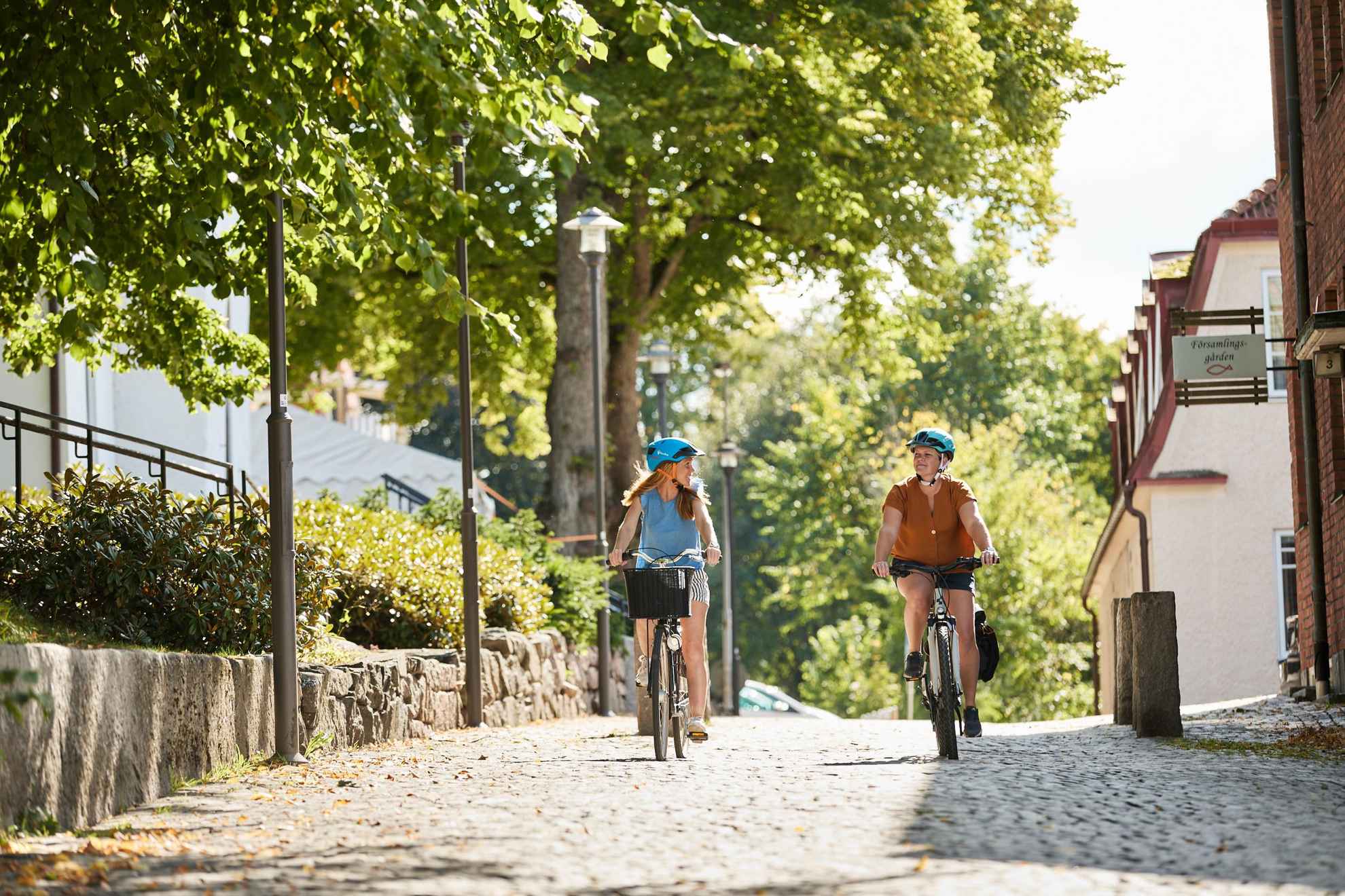 Deux femmes font du vélo dans une petite rue pavée. Maisons et verdure le long de la rue.