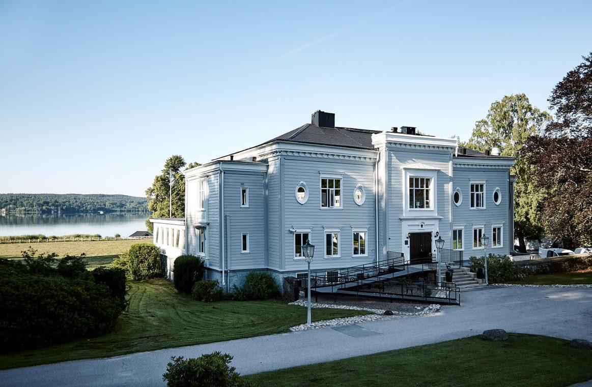 Vue sur le manoir d'Aspenäs, une grande maison en bois gris. Le manoir est entouré de verdure et est situé au bord du lac Aspen.