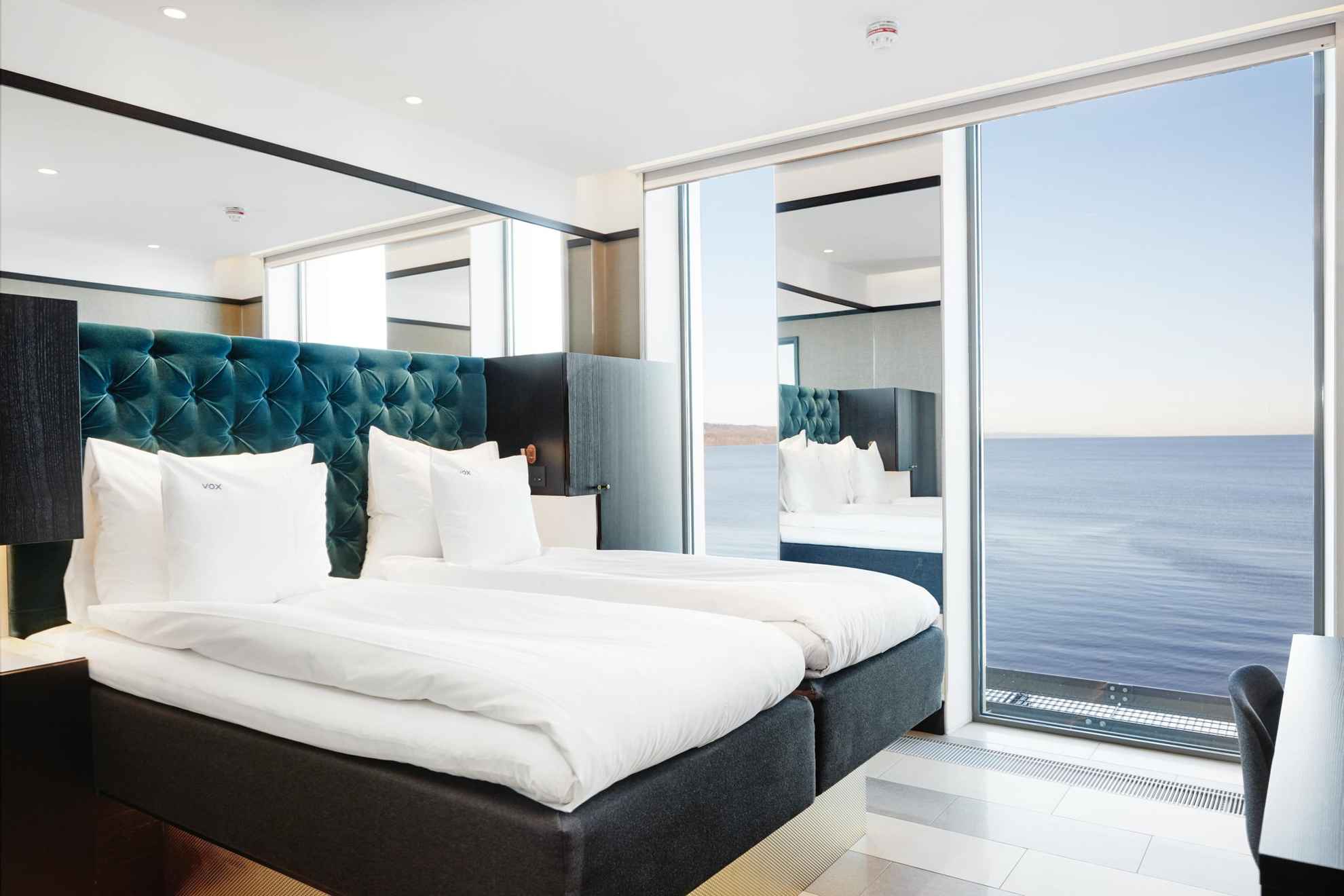 Une chambre d'hôtel avec un lit double et des draps blancs. La chambre possède de grandes ouvertures qui donnent sur le lac Vättern.