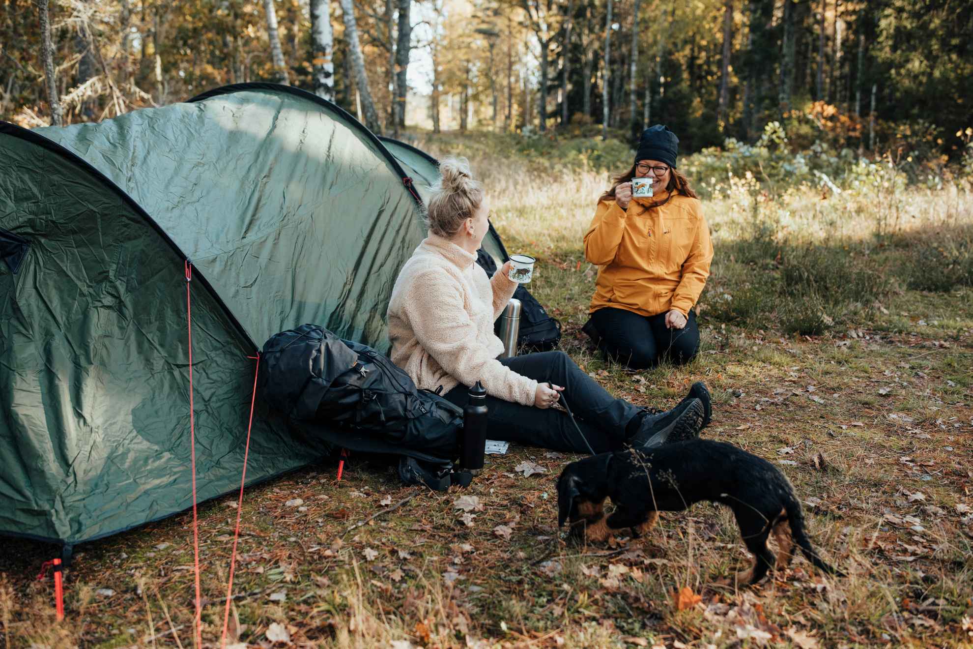 Deux femmes vêtues de vêtements de randonnée sont assises par terre à côté d'une tente et boivent du café dans des tasses. Au premier plan, on voit un teckel noir.