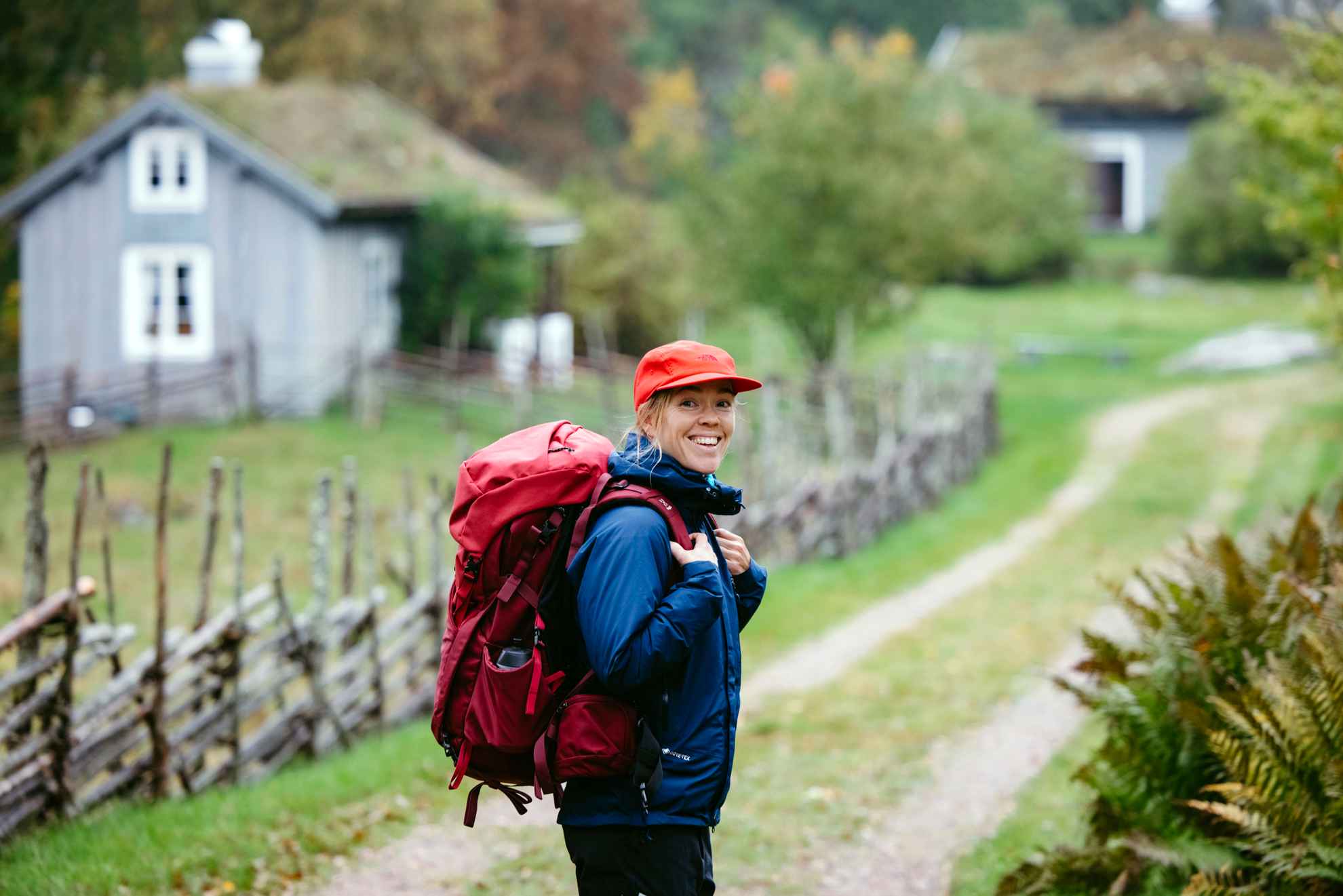 Une femme portant des vêtements de randonnée et un sac à dos regarde la caméra depuis un sentier de randonnée. En arrière-plan, on aperçoit une ferme en bois.