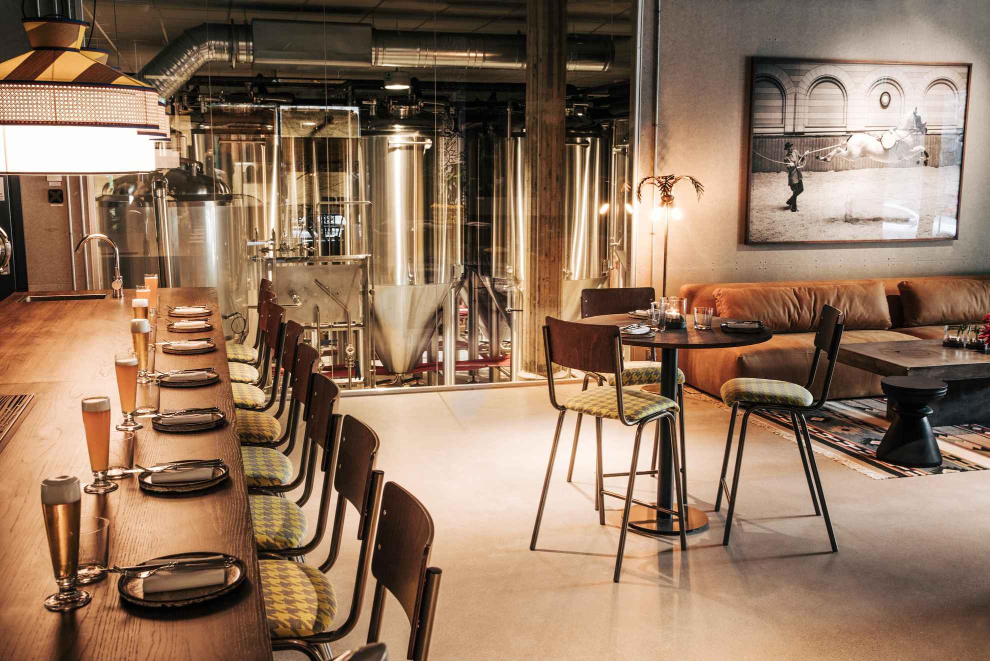 Inside a restaurant, the bar to the left is set with glasses of beer, there is a set table with three chairs and a sofa to the right. In the background behind a glass wall you can see a brewery.