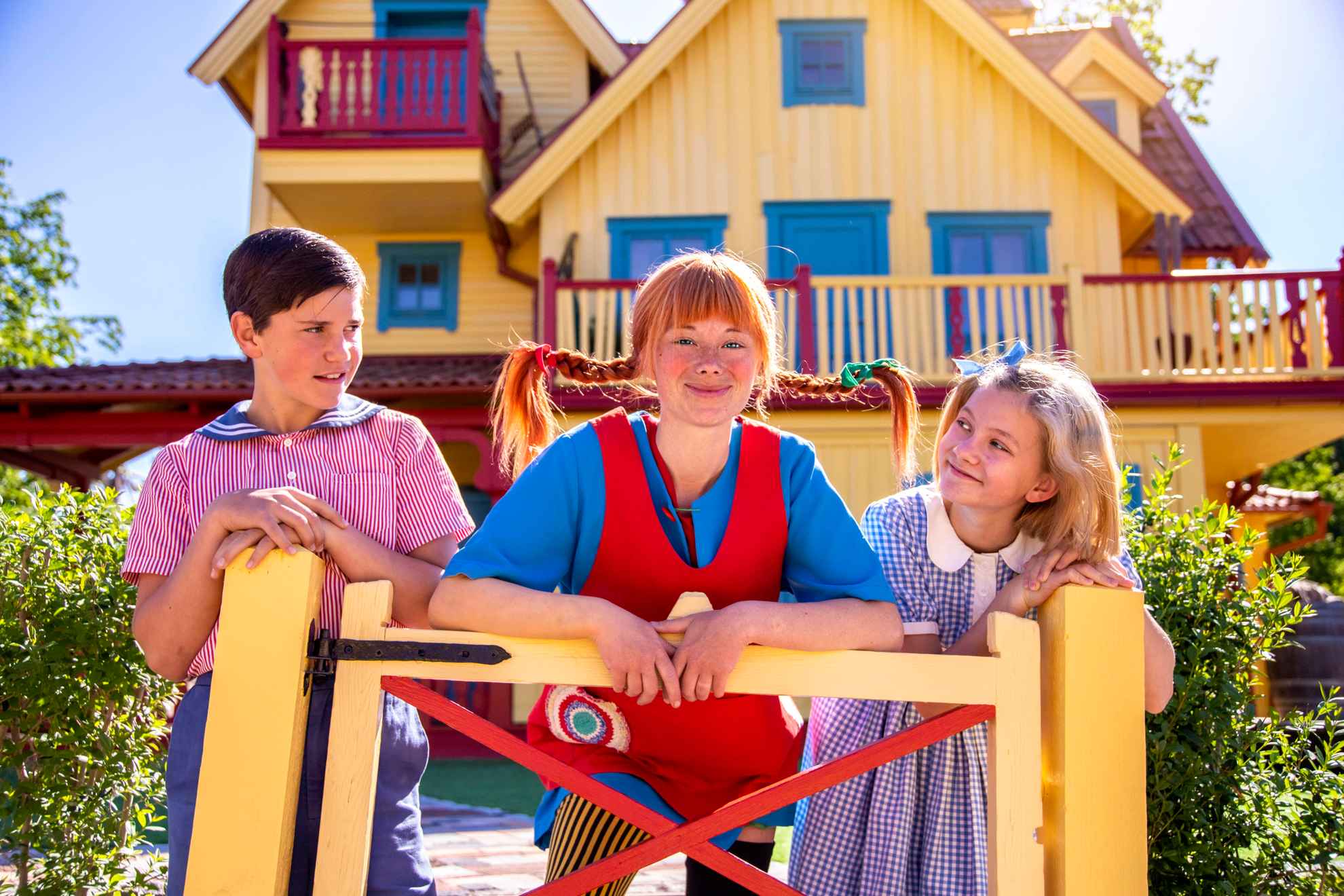 Des acteurs déguisés en Tommy, Fifi et Annika se tiennent près d'un portail jaune devant une maison jaune (Villa Villekulla).