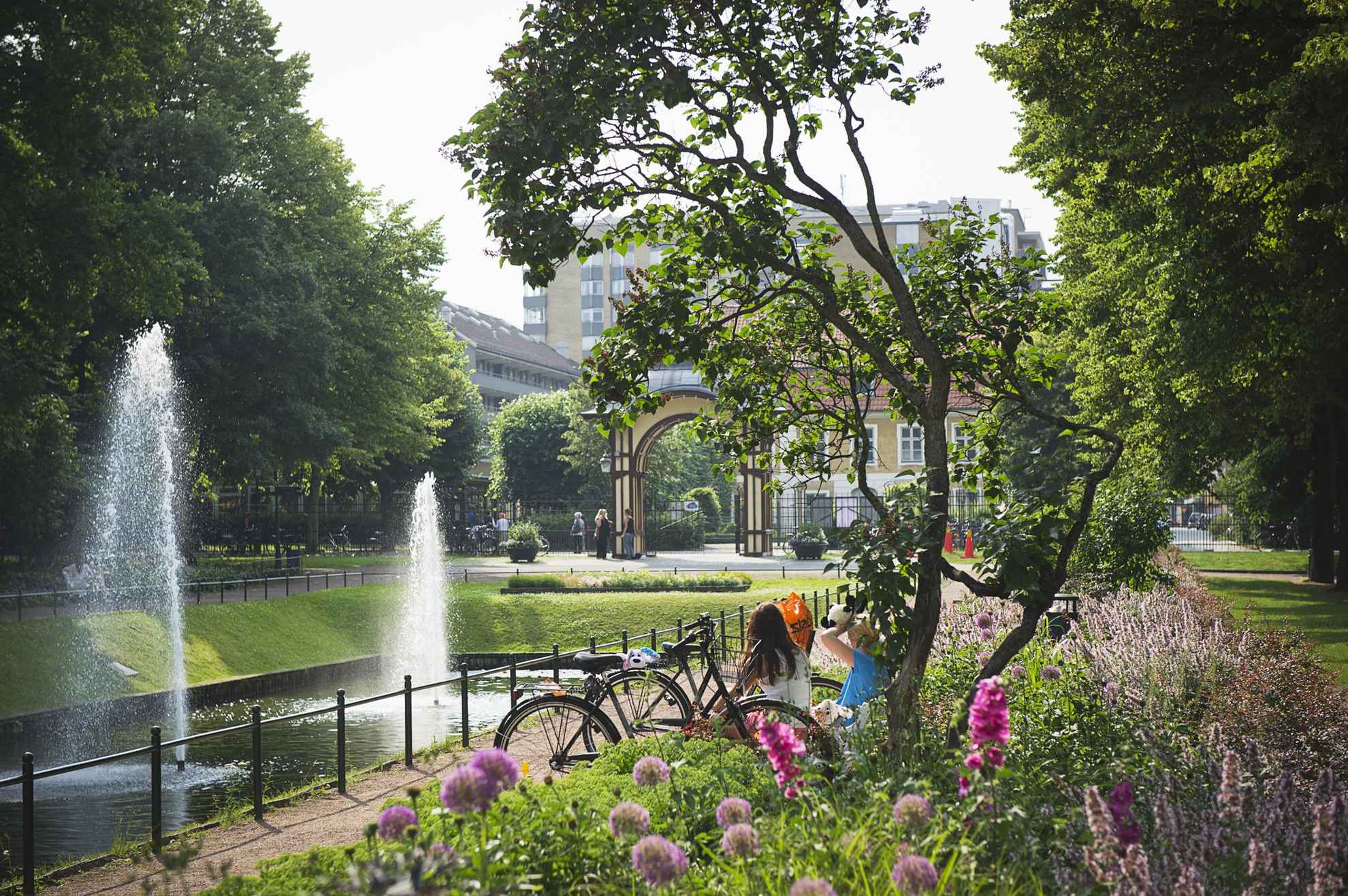 Le parc est rempli de fleurs et de verdure. On aperçoit le dos de deux femmes assises à côté d'un plan d'eau avec deux fontaines. L'entrée du parc se trouve en arrière-plan et on distingue des bâtiments derrière le parc.