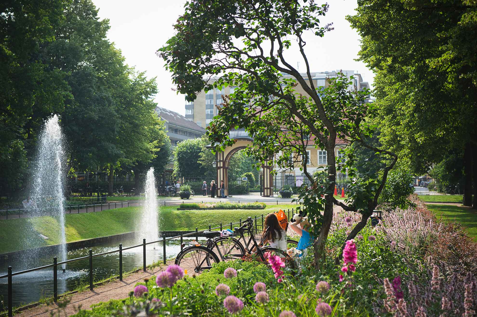 Fleurs et verdure à l'intérieur d'un parc urbain. On voit le dos de deux femmes assises au bord d'un bassin avec deux fontaines. À l'arrière-plan, on voit l'entrée du parc et quelques immeubles à l'extérieur du parc.