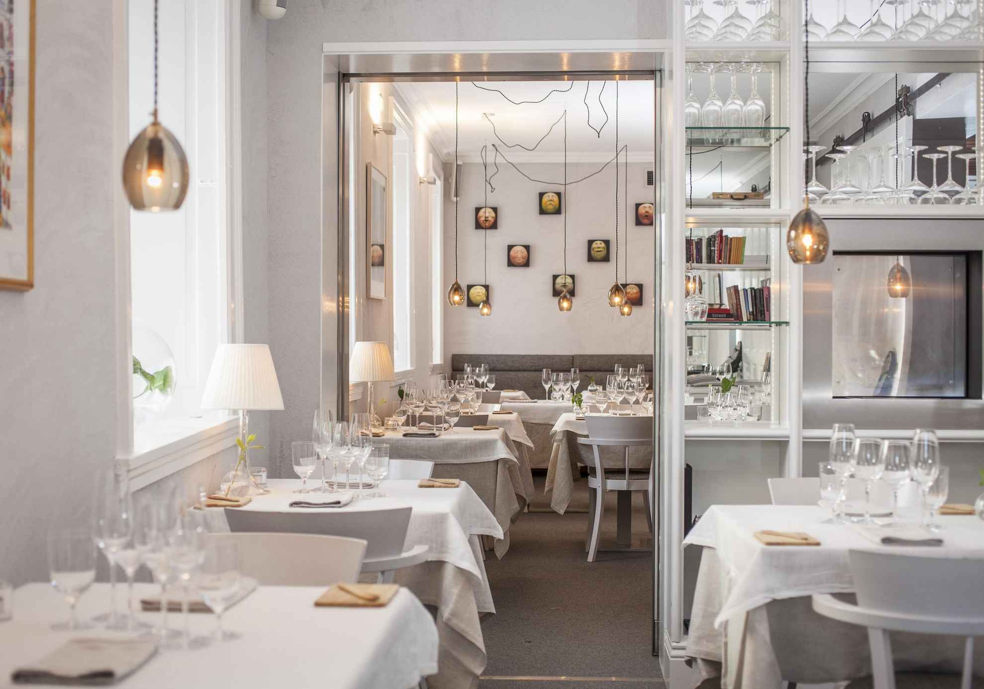 Dans un restaurant aux murs blancs, les tables sont dressées avec des nappes blanches, des serviettes en lin et des verres à vin. Des lampes sont suspendues au plafond.