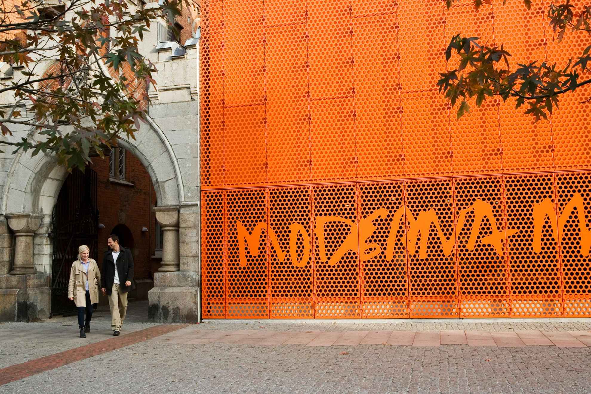 Un homme et une femme marchant dehors et passant devant le signe orange du musée Moderna Museet de Malmö.