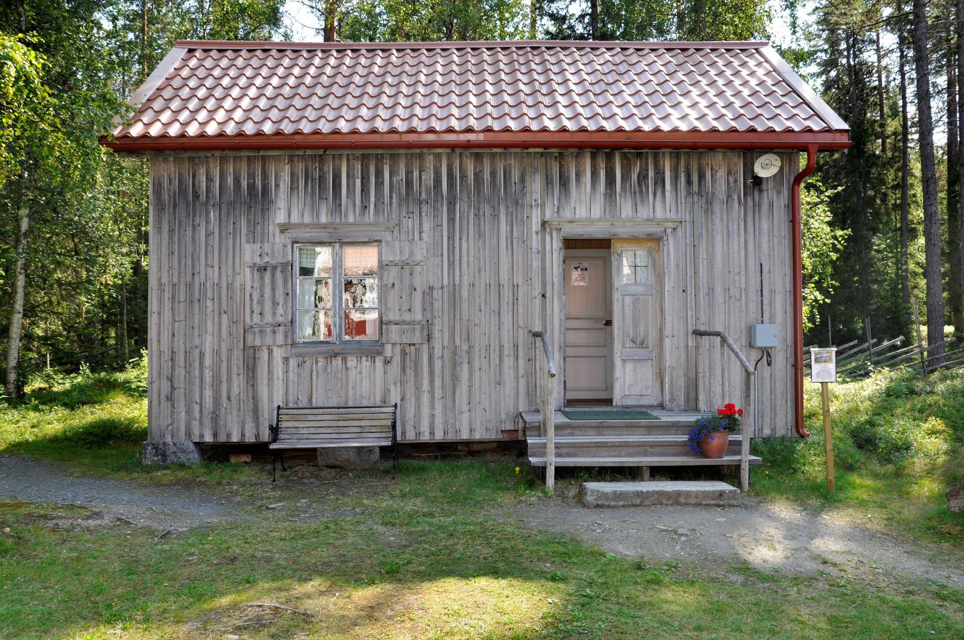 Une maison en bois gris avec un pot de fleurs à l'entrée, est situé en face d'une forêt.