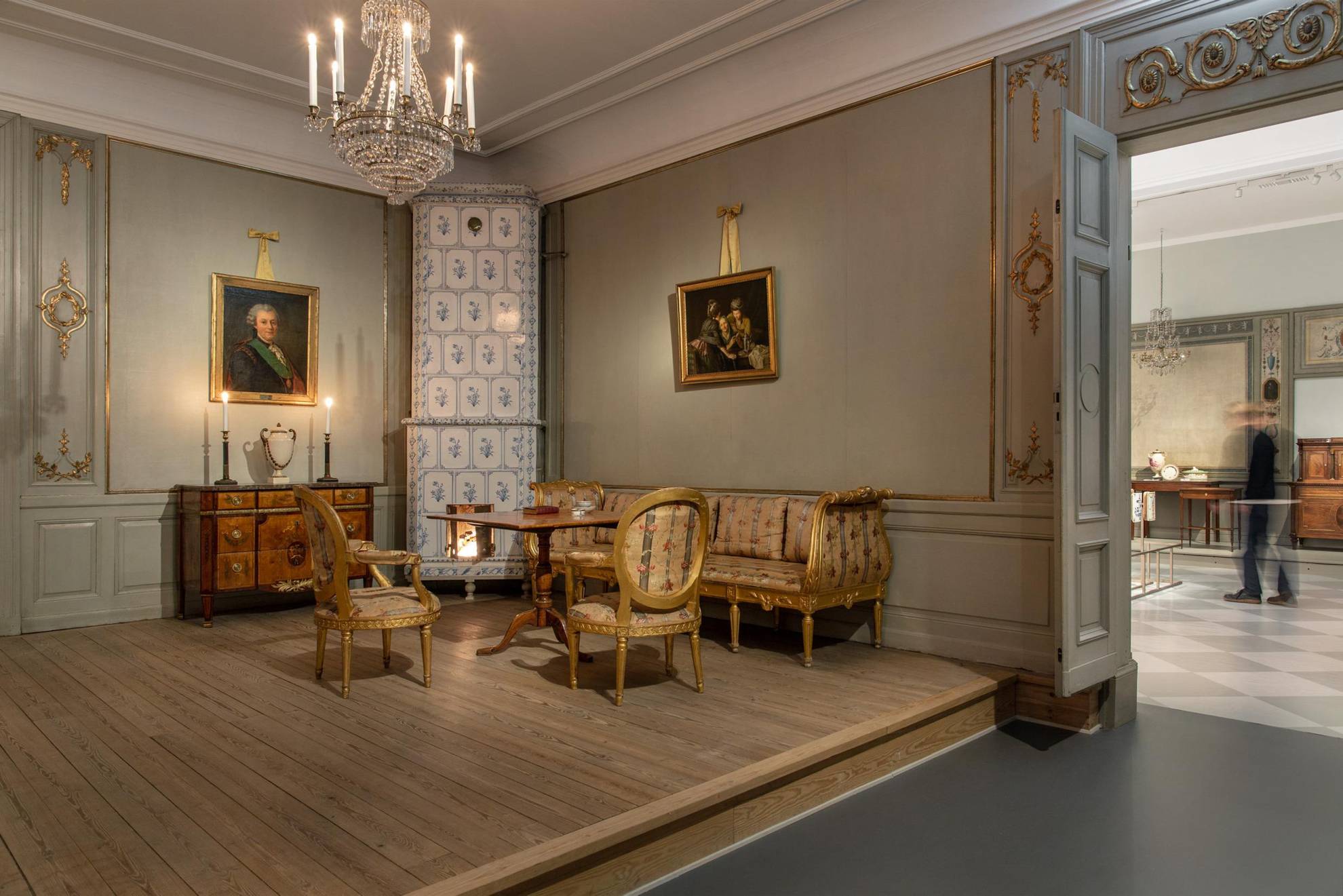 Mobilier du 18e siècle dans une exposition. On voit sur la photo un poêle en faïence, un canapé et deux chaises, une table et une commode, un lustre en cristal suspendu au plafond et deux tableaux au mur.