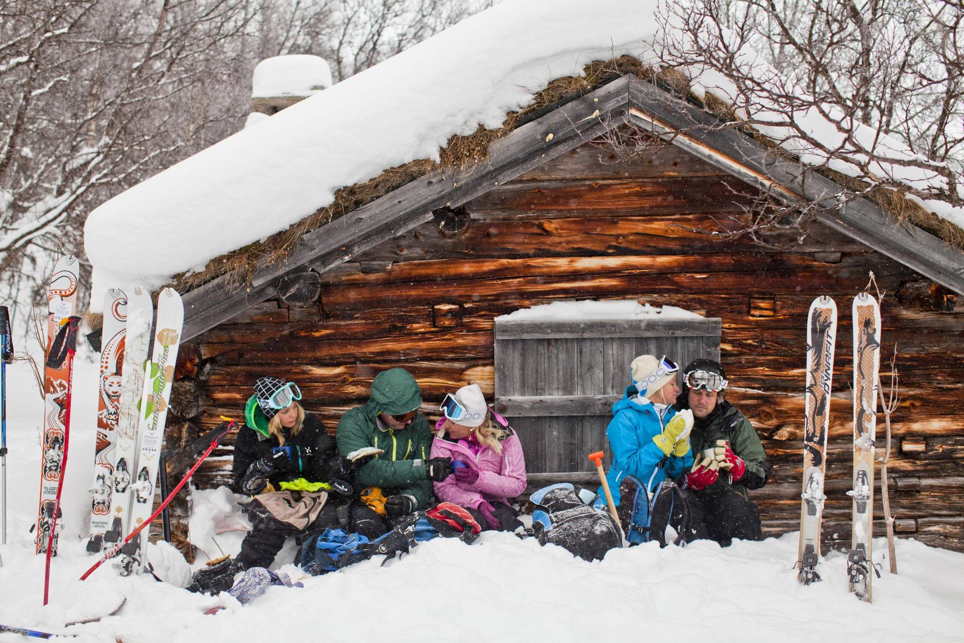 Cinq personnes sont assises dans la neige devant une maison en bois dans les montagnes et font une pause café après avoir skié.