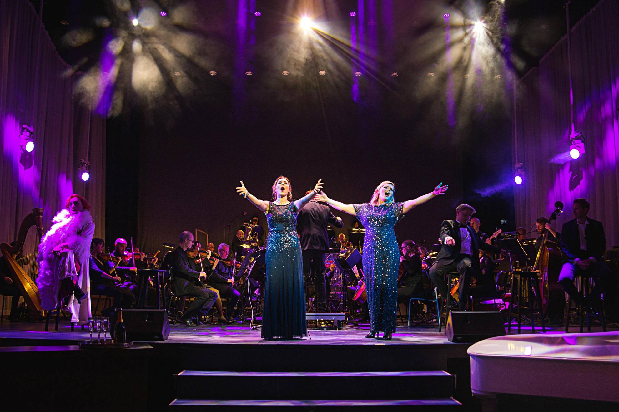 Deux femmes dans des robes bleues à paillettes sur une scène lors d'un spectacle. Il y a un orchestre derrière eux.