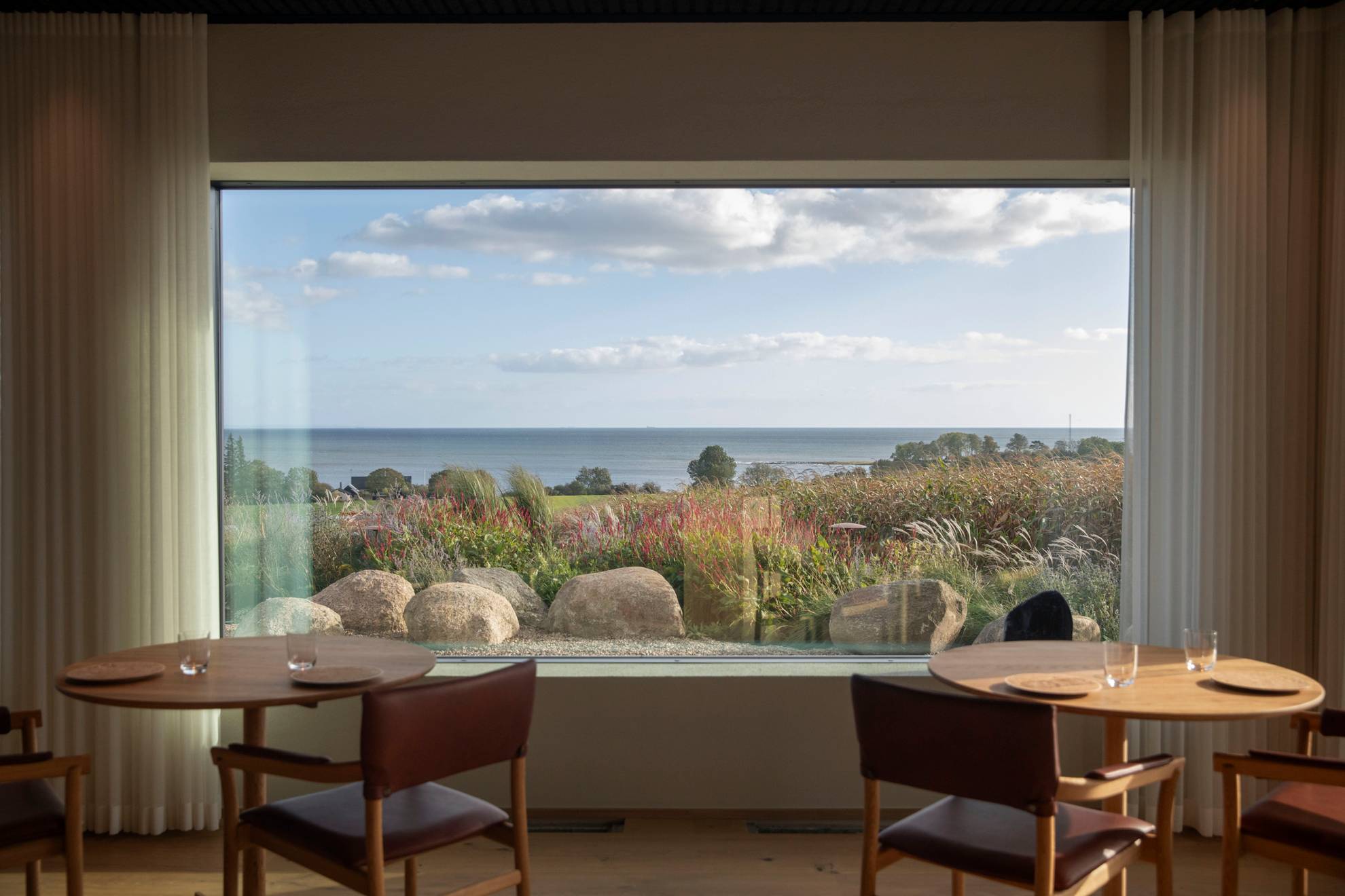 Two tables with chairs at Vyn restaurant, standing in front of a large window and overlooking the sea on a summer day.