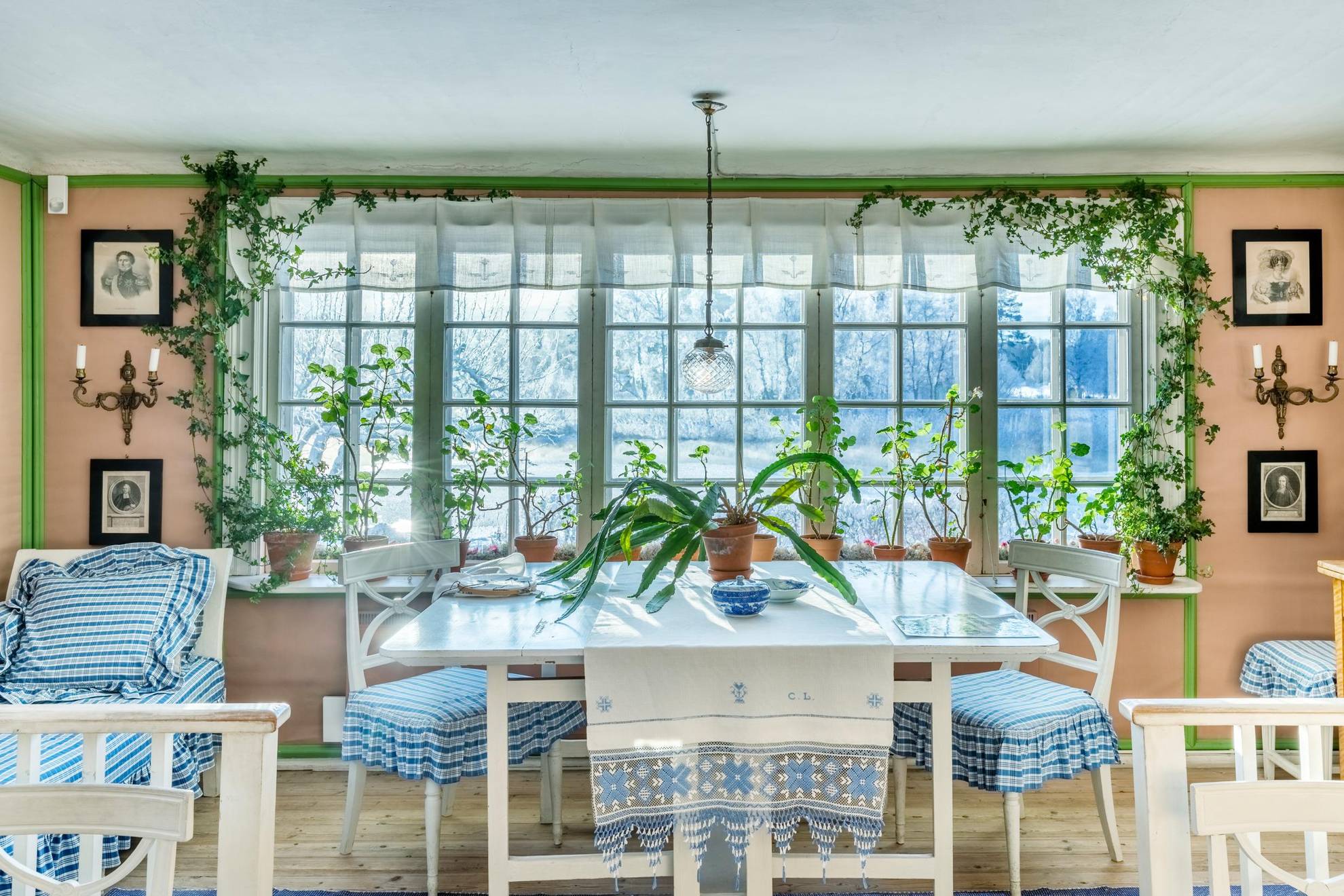 Une cuisine avec une grande fenêtre, de nombreuses plantes et des tissus blancs et bleus.