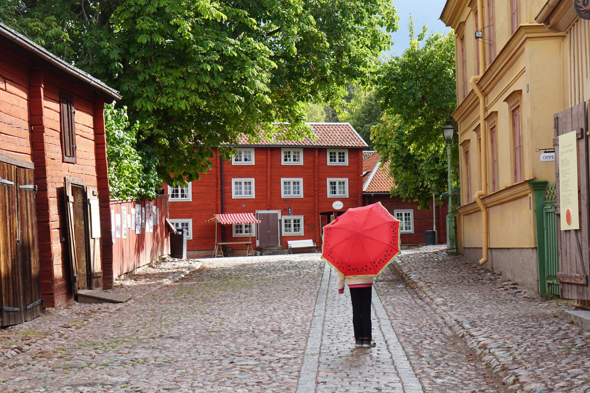 Une personne marche avec un parapluie rouge dans une rue pavée entre de vieilles maisons.