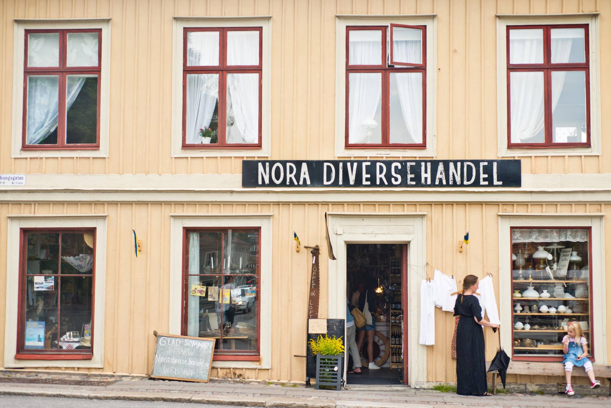 Une petite boutique abritée dans une maison en bois jaune dans une rue de la vieille ville. Une pancarte au-dessus de la boutique indique "Nora Diversehandel". Une femme se tient debout et une petite fille est assise sur un banc devant la maison.