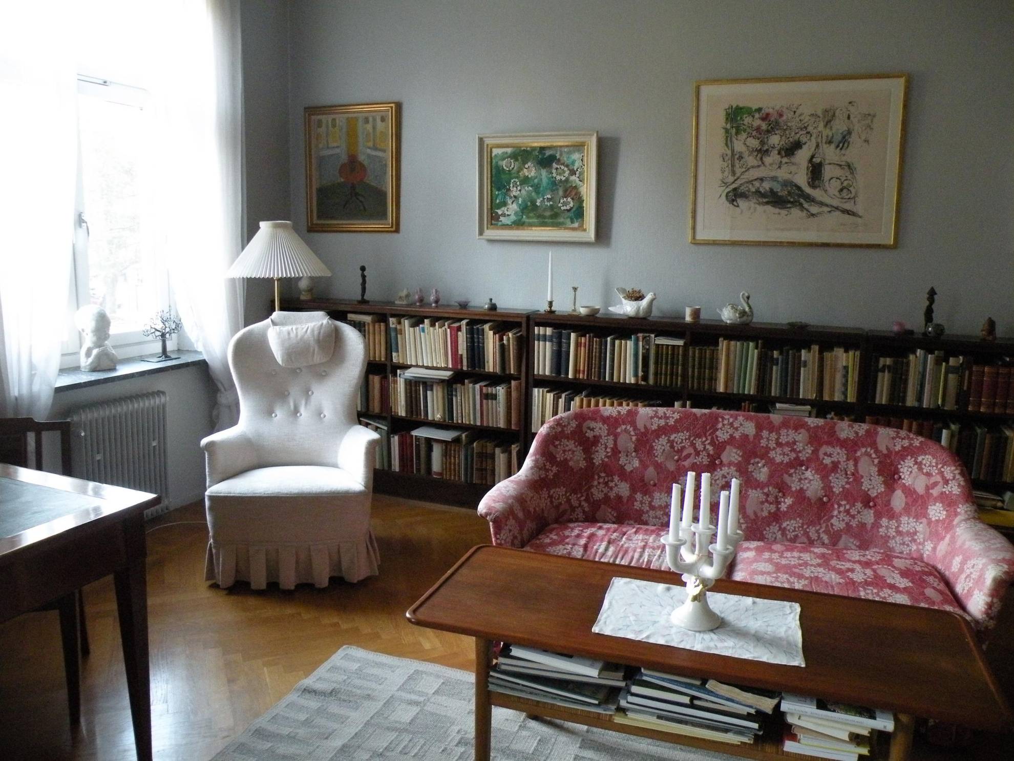 Le salon d'Astrid Lindgren, avec une chaise pour lire, une grande bibliothèque sur laquelle sont posés des bibelots, un canapé rouge à motifs et quelques peintures au mur.